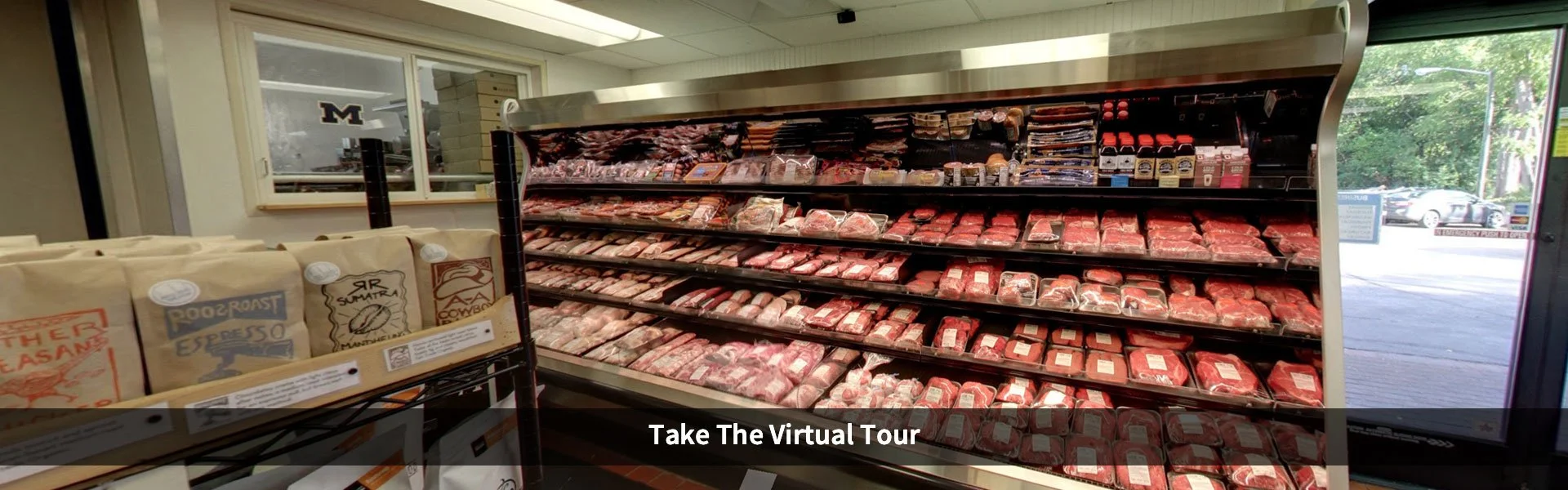 Refrigerated meat display case with various packaged cuts of beef in a grocery store, with a window in the background showing trees outside.
