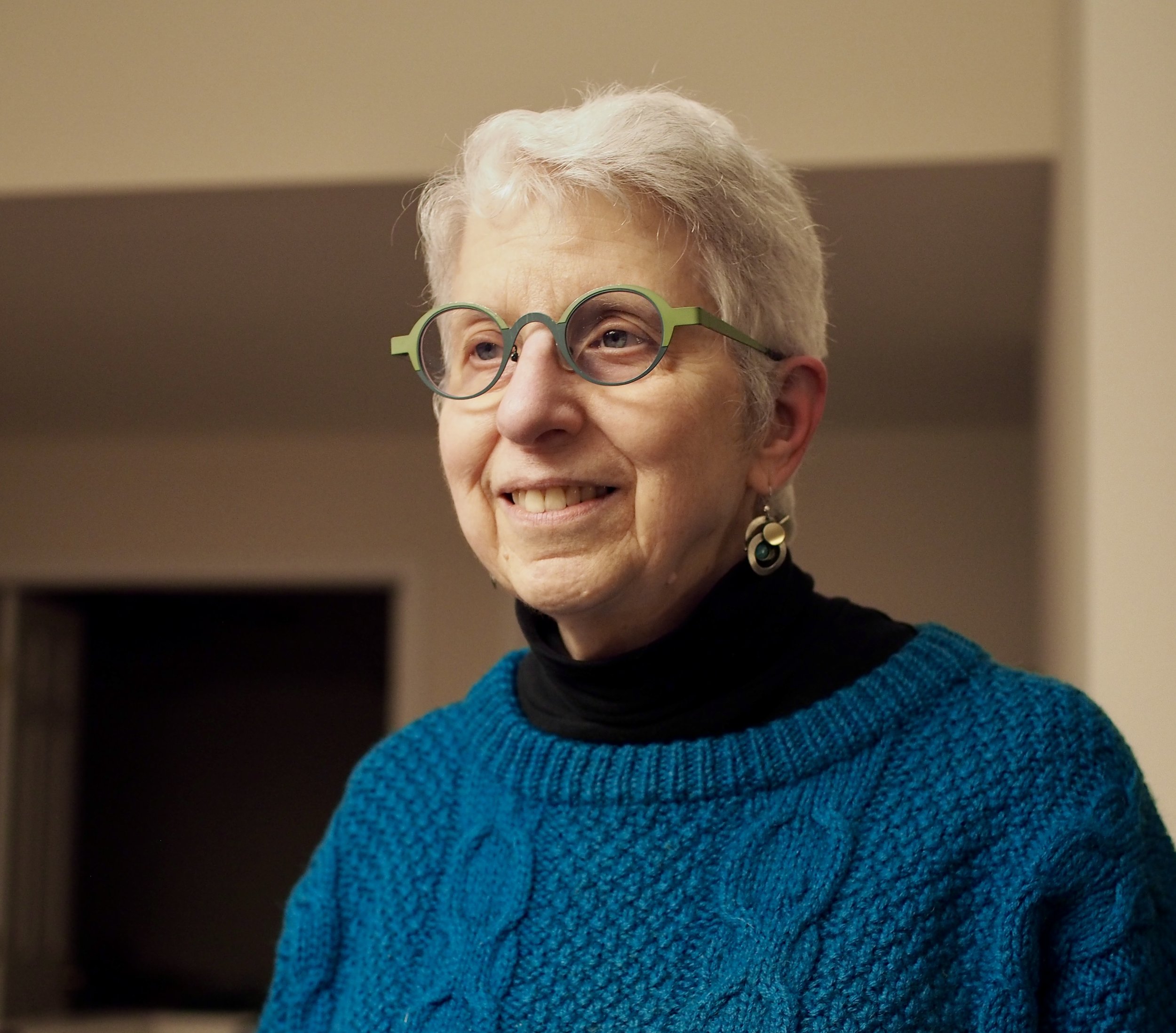 A woman with short gray hair wearing green-rimmed glasses, a black turtleneck, and a blue knitted sweater with a textured pattern. She is smiling and wearing dangling earrings.