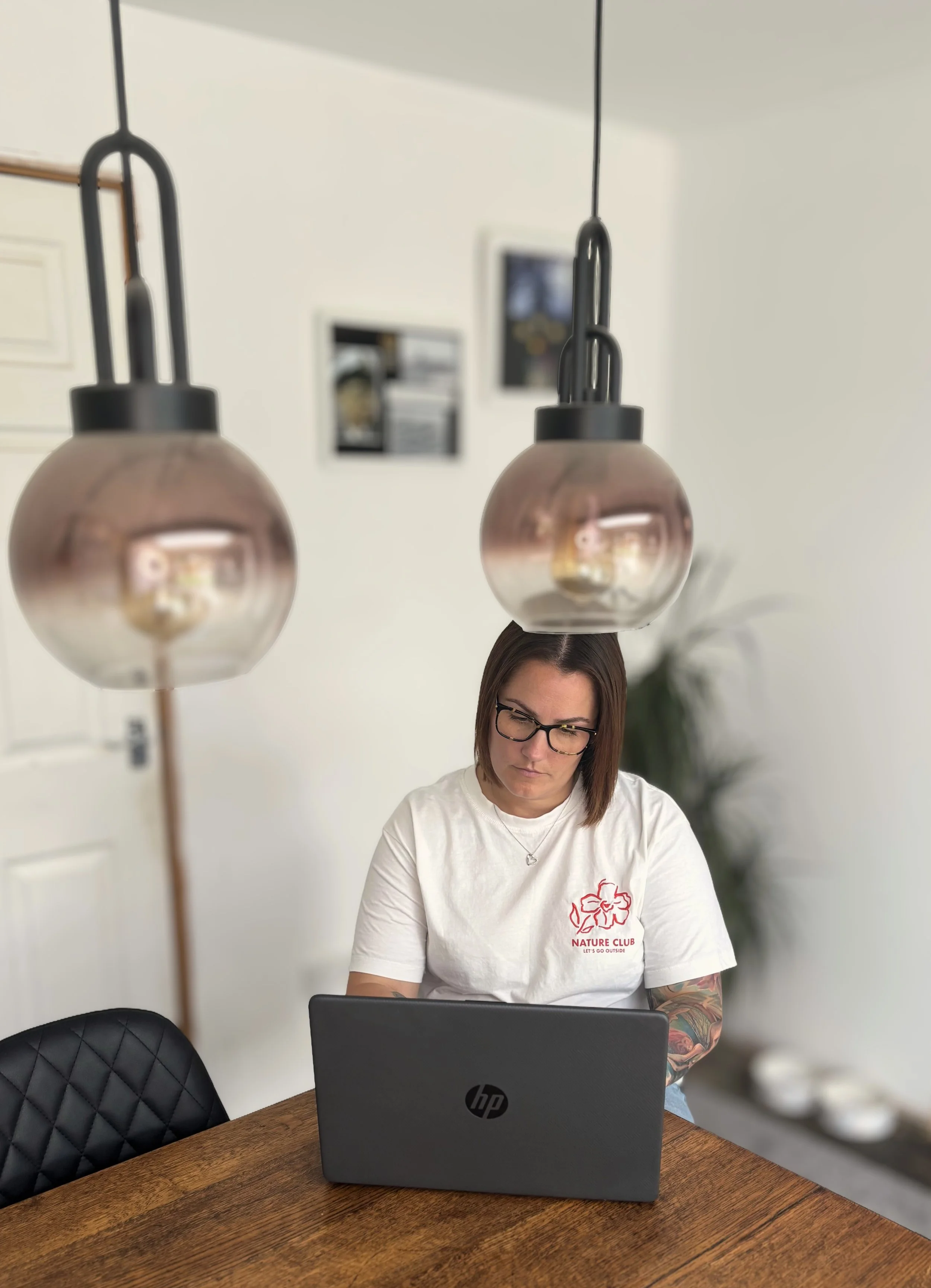 A woman working on a laptop at a wooden table, viewed through two hanging pendant lights, with a blurred background of a white wall and some framed pictures.