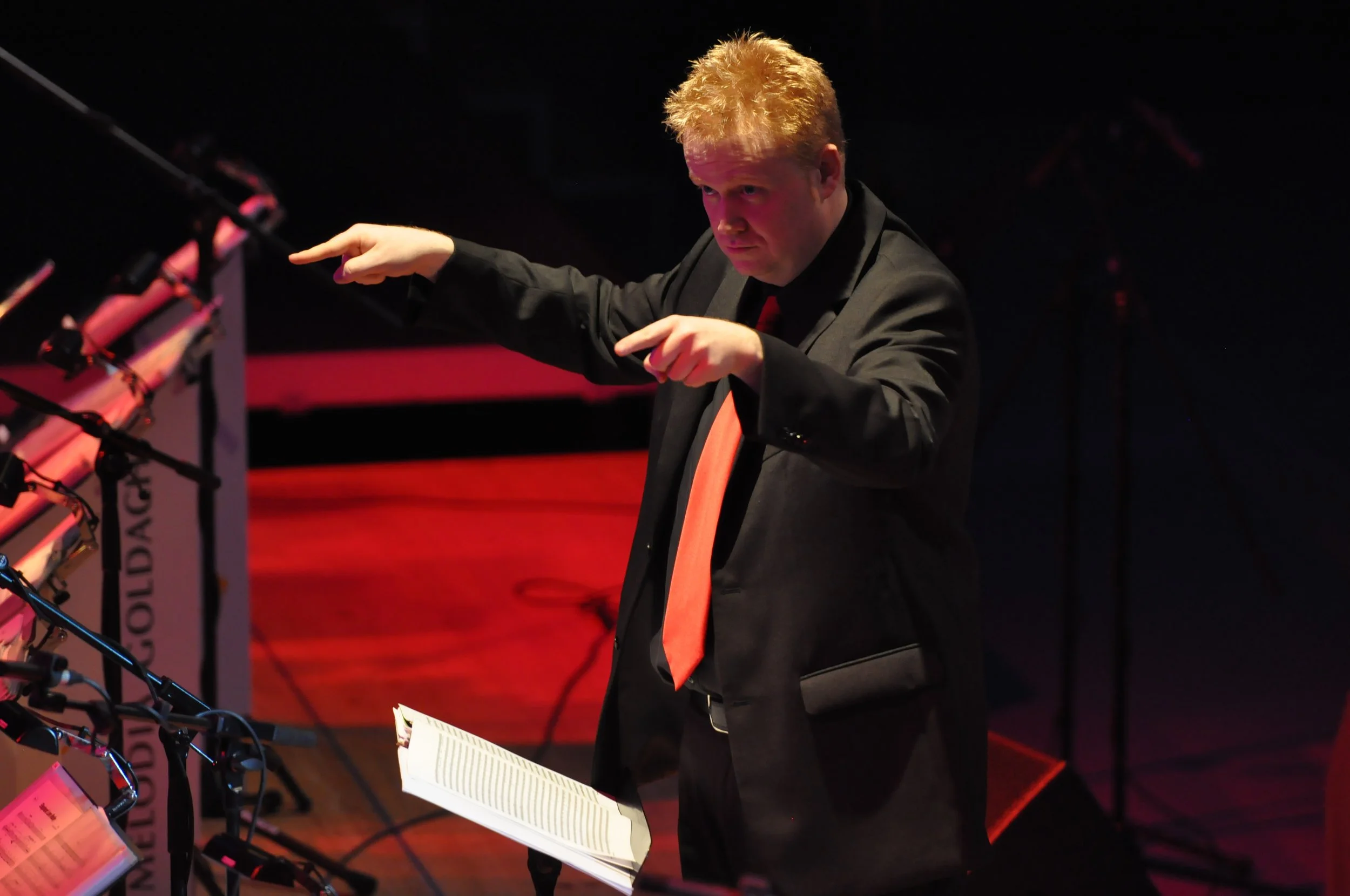 Ein Mann in dunklem Anzug dirigiert ein Orchester auf einer Bühne, mit Notenständer vor ihm und Mikrofonen auf der linken Seite.