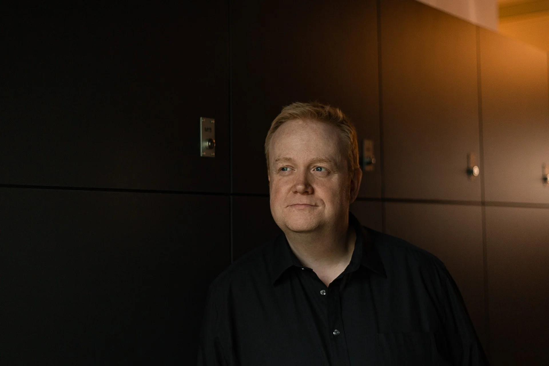 Ein Mann mit rotblonden Haaren in dunkler Kleidung lehnt an einer dunklen Wand in einem Gebäude, Blick nach rechts, leicht nach oben gerichtet, leichter Sonnenuntergangs- oder Innenraumlicht, ruhige Stimmung.