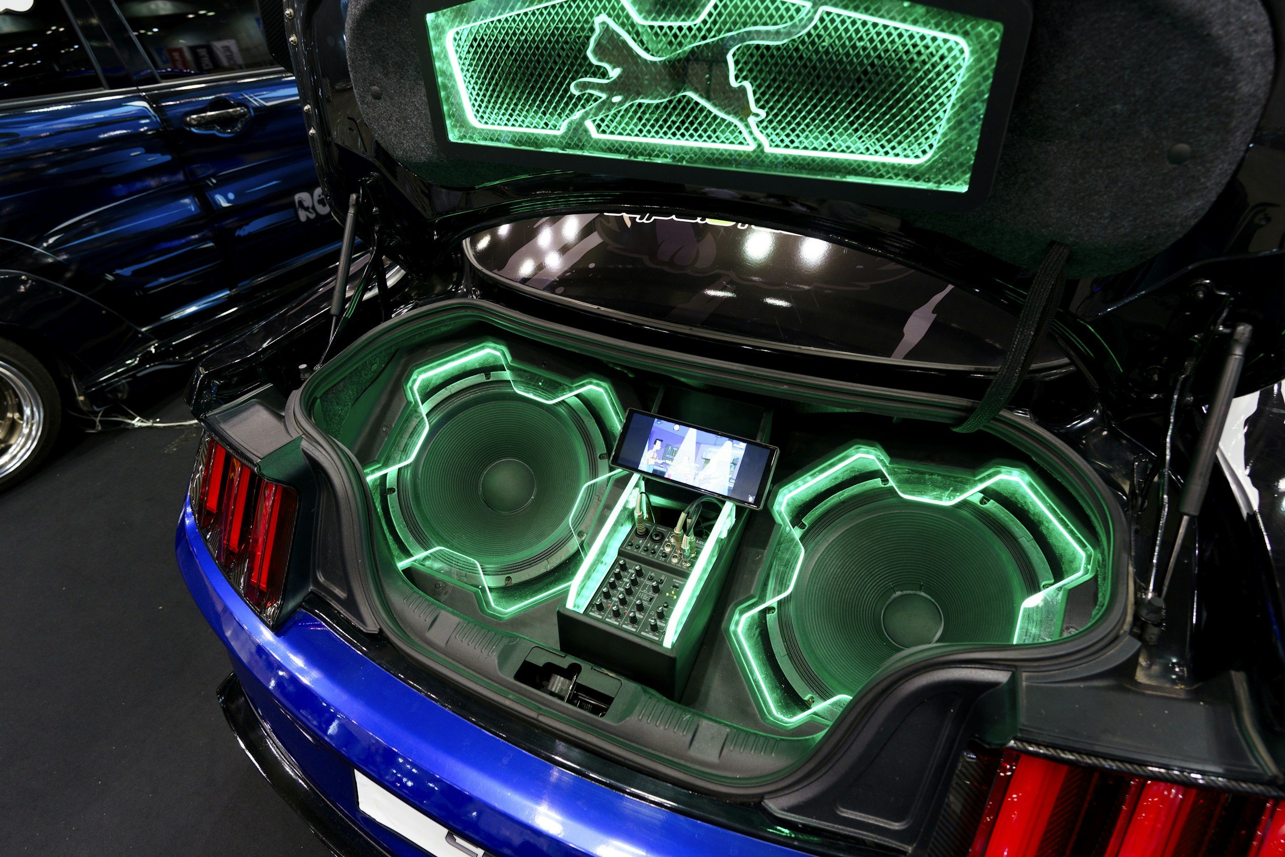 The image shows the open trunk of a blue car equipped with large green speakers, two illuminated neon signs with the Red Bull logo, and a smartphone mounted on a stand displaying a digital control interface.