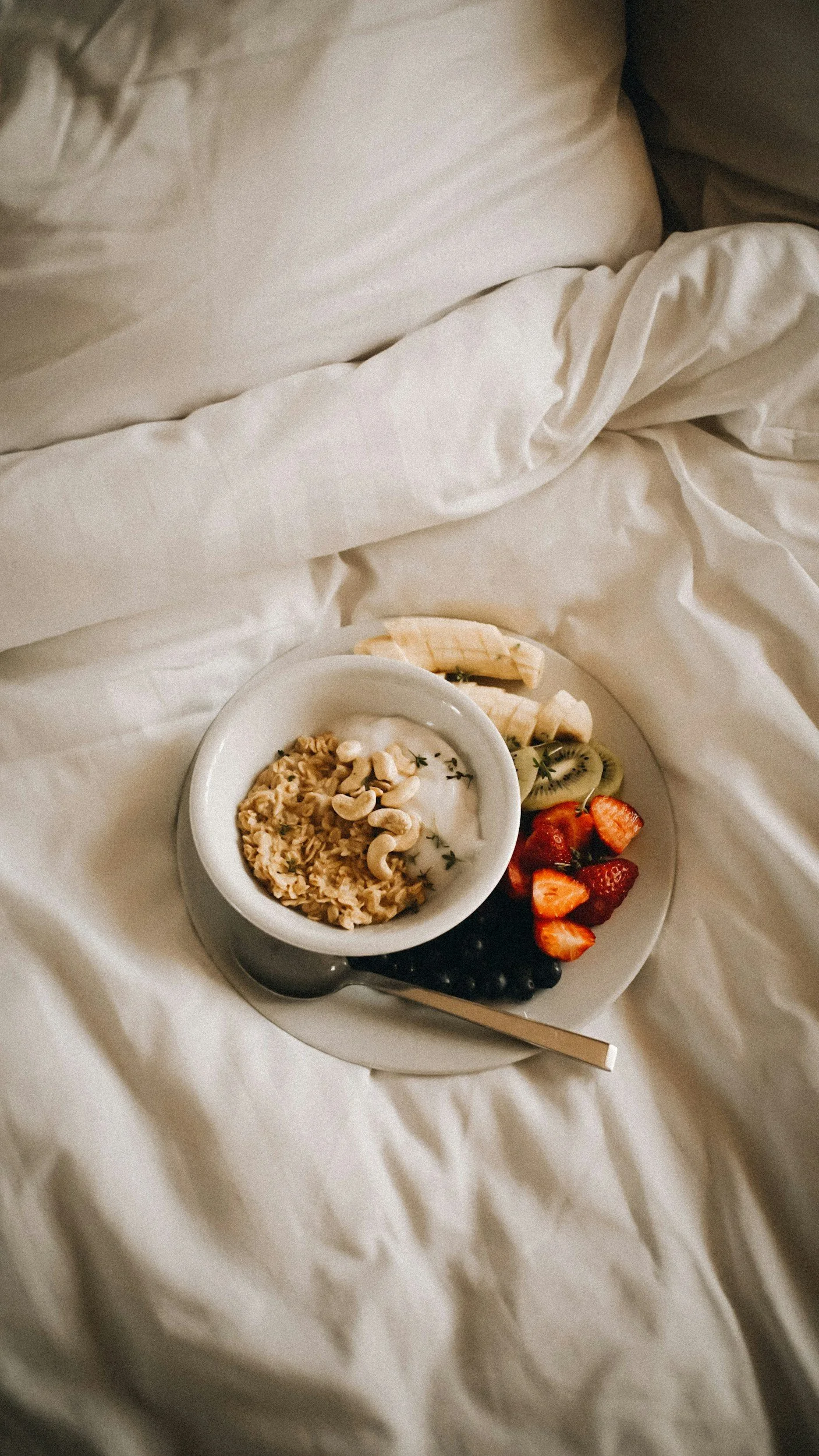 Breakfast tray with cereal, yogurt, banana, kiwi, strawberries, blueberries, and cashews on a bed with white sheets.
