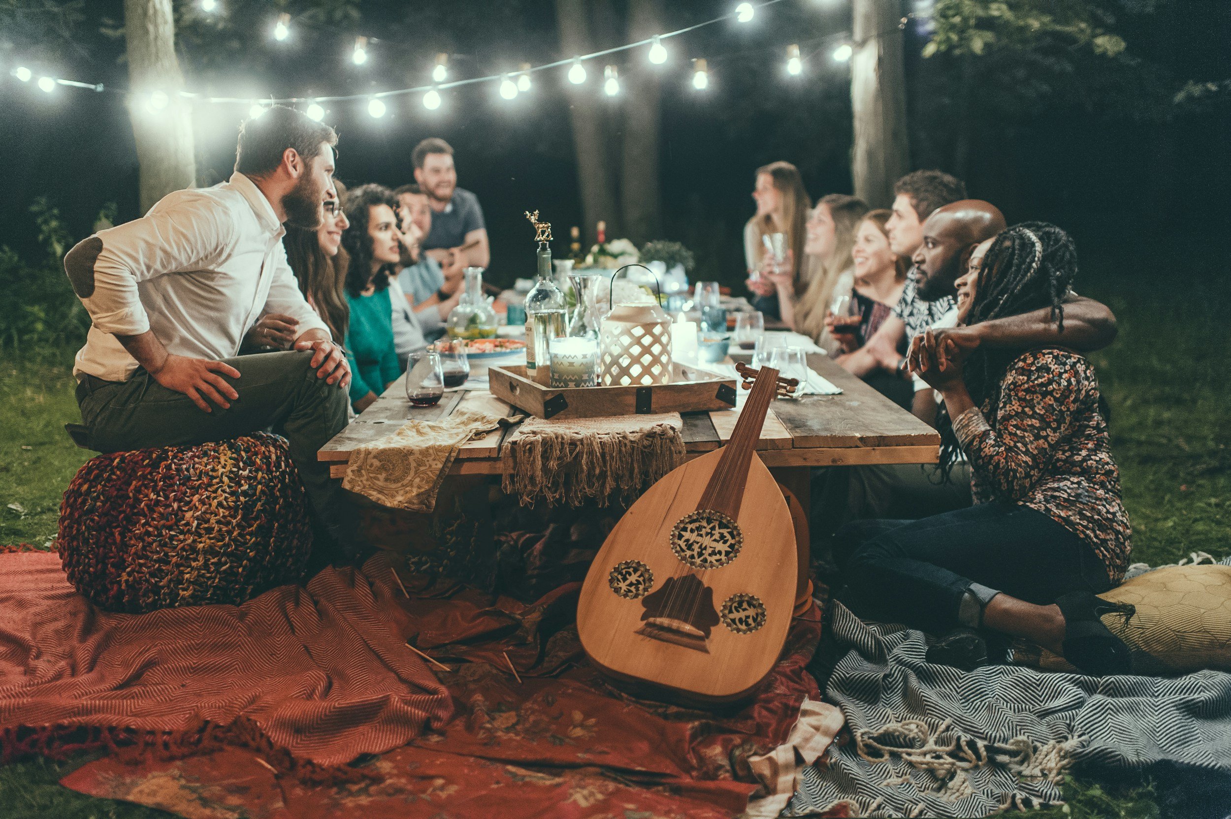 People gathered around a wooden outdoor table for a nighttime dinner, decorated with string lights, candles, and lanterns, with a guitar placed on a blanket in the foreground.