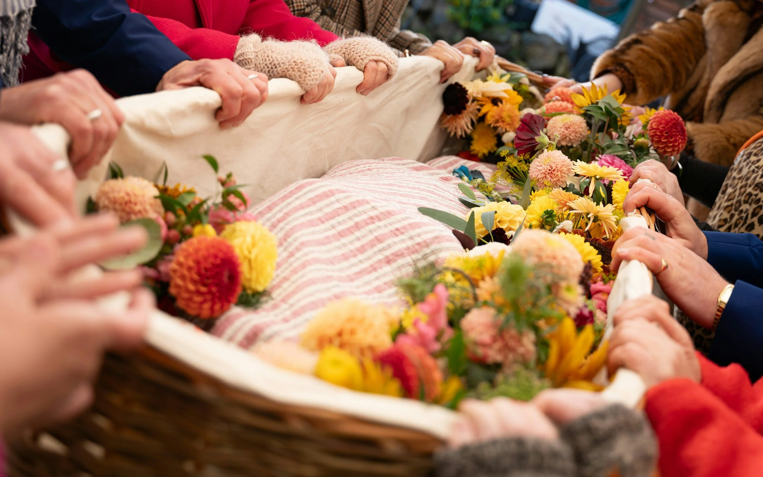 People holding a wicker basket decorated with flowers and a pink and white striped cloth, likely part of a funeral or memorial service.
