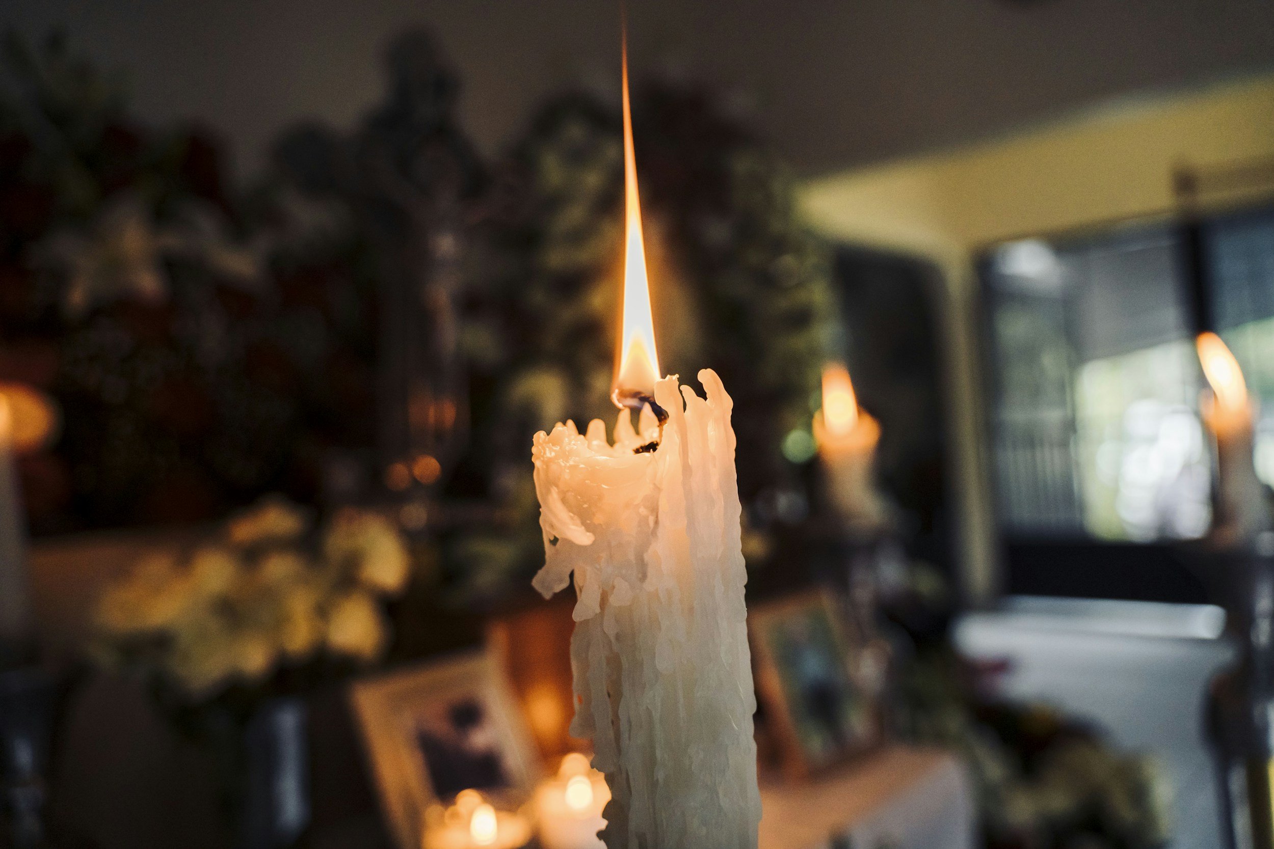 Close-up of a single burning white candle with melted wax dripping down, in a dimly lit room with blurred background candles and picture frames.