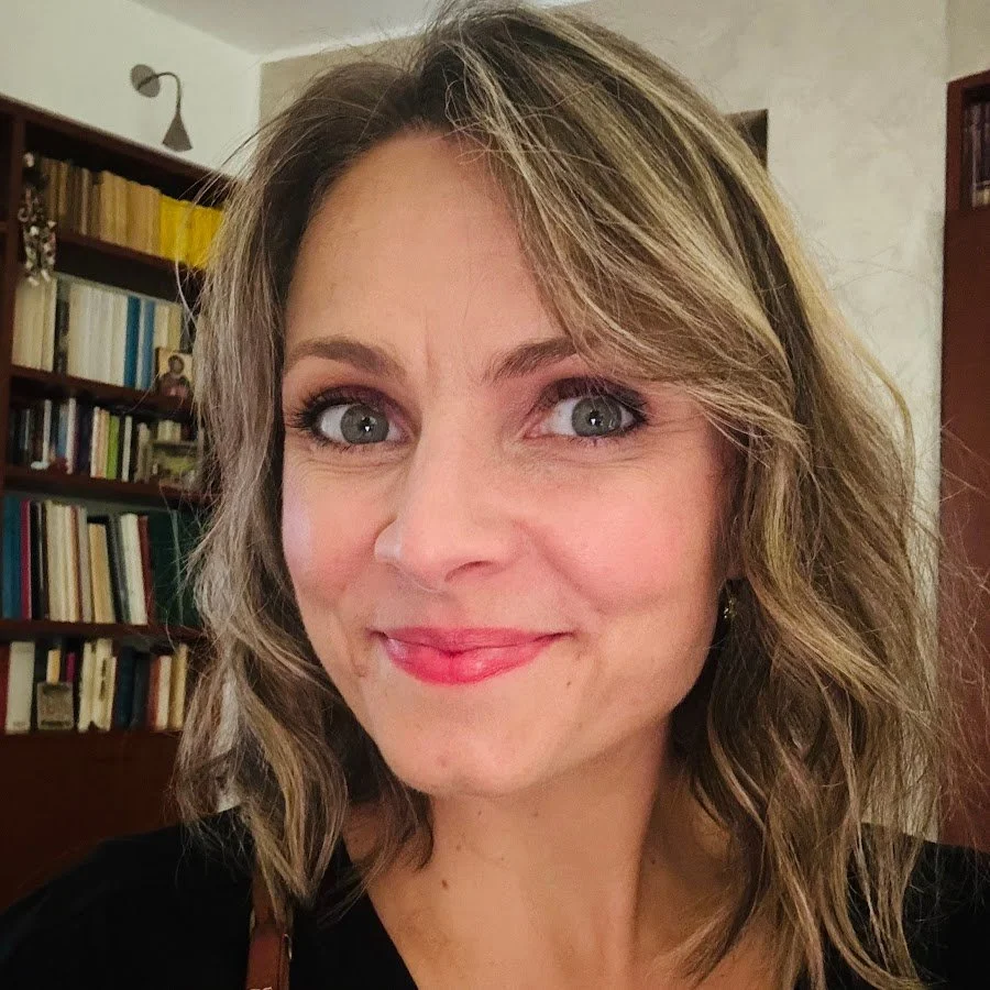 A woman with shoulder-length wavy blonde hair and blue eyes, smiling in front of a bookshelf filled with books. Katia di Munno Summer Masterclass Bergamo