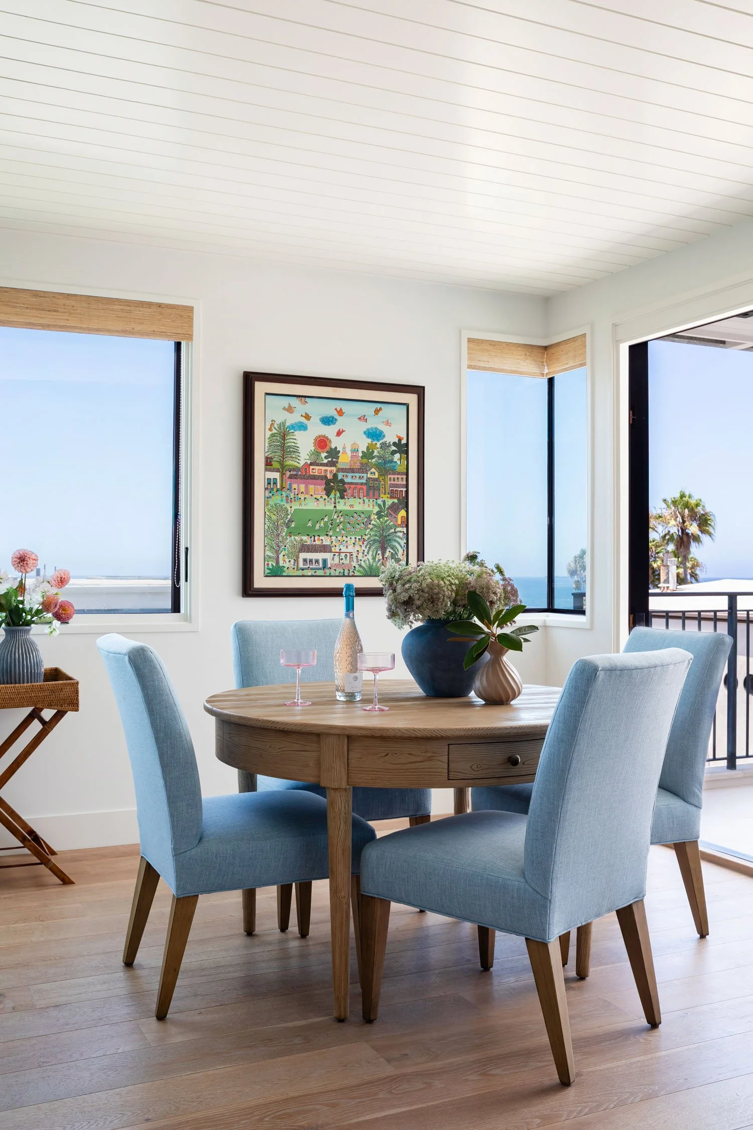 Coastal dining room with round wood table, blue upholstered chairs, colorful artwork, and ocean views in Manhattan Beach, designed by JKB Home Design