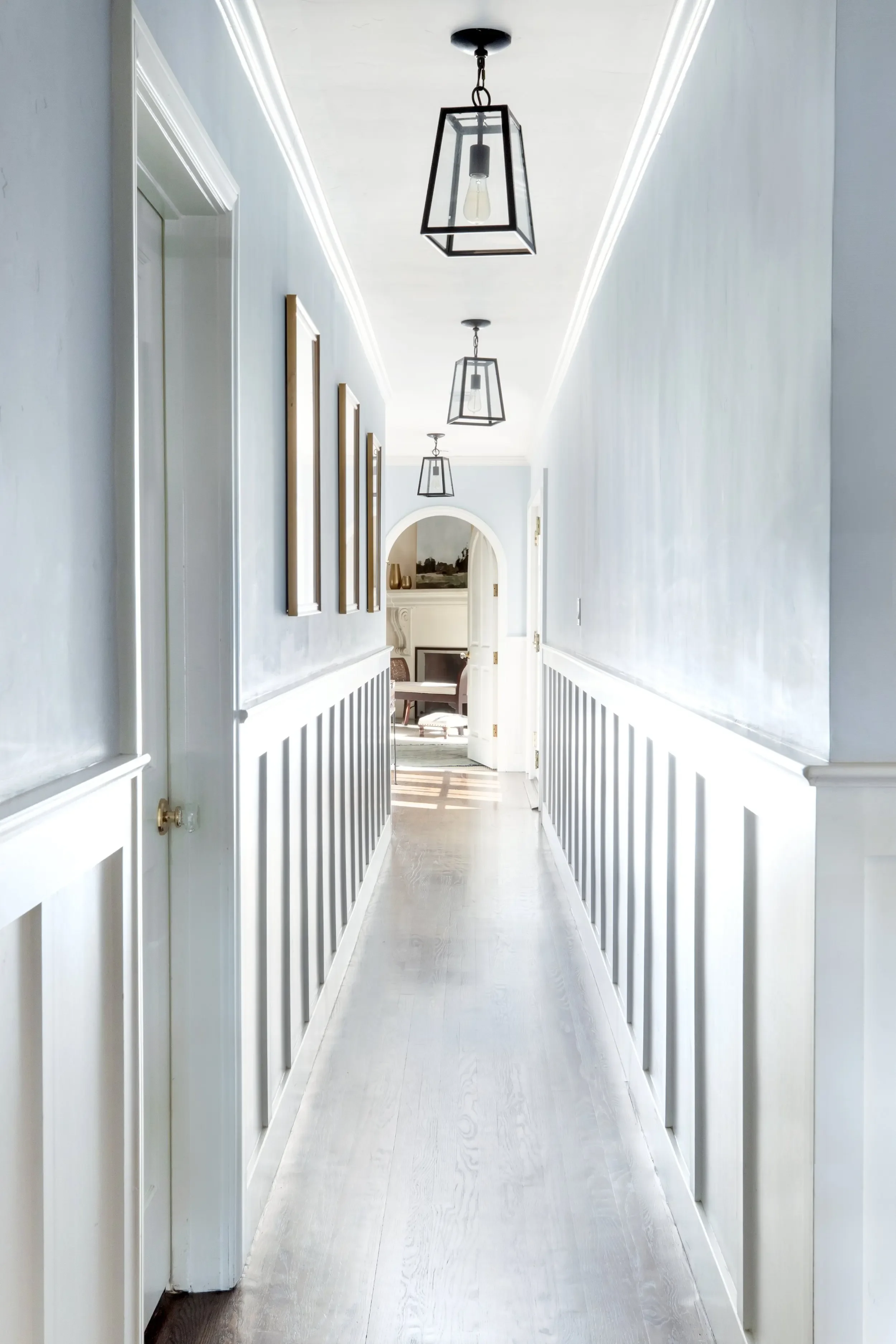 Pale blue hallway with white wainscoting, wood floors, and black lantern pendants in Palos Verdes Estates, designed by Julie Beuerlein