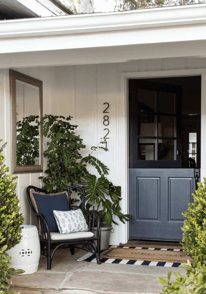 Covered front entry with wall mirror, woven chair, layered doormats, and lush greenery