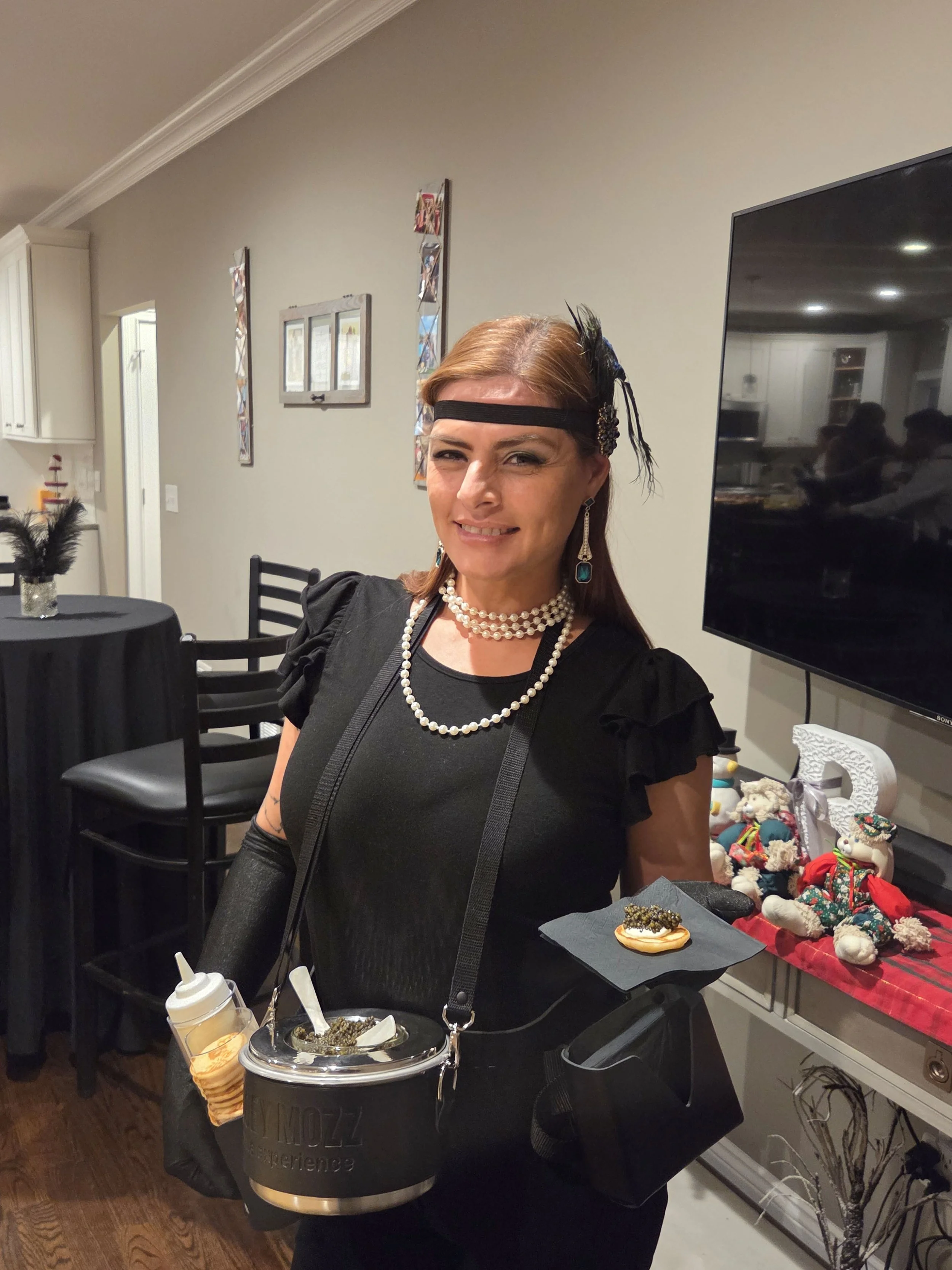 Caviar Catering Service in vintage 1920s flapper style costume with headband, layered pearl necklaces, black dress, and long gloves, standing in a living room with holiday decorations, holding a treat and a container of caviar.