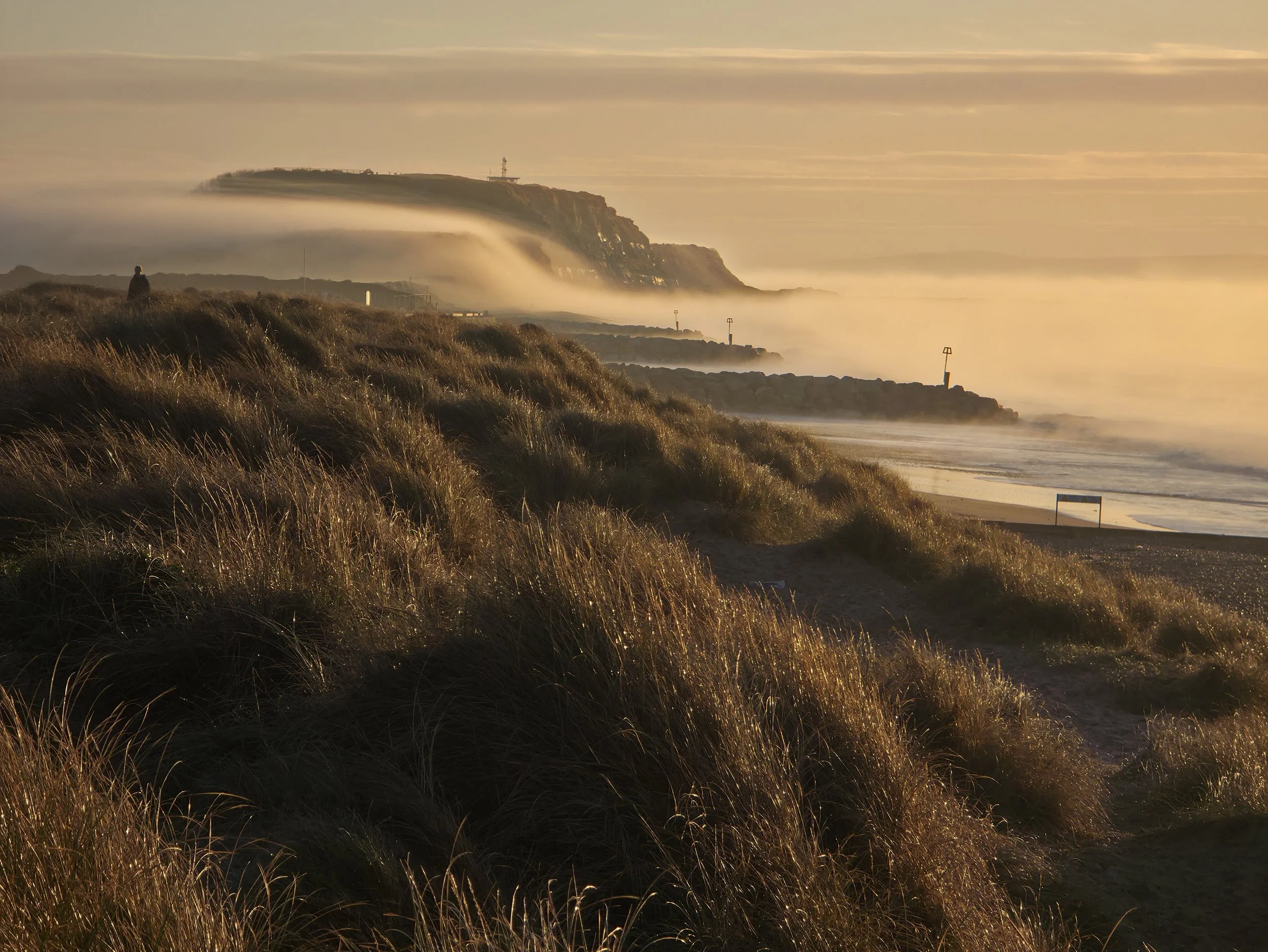 A person standing on a grassy cliff overlooking the ocean at sunset, with fog and mist over the water and a lighthouse in the background.