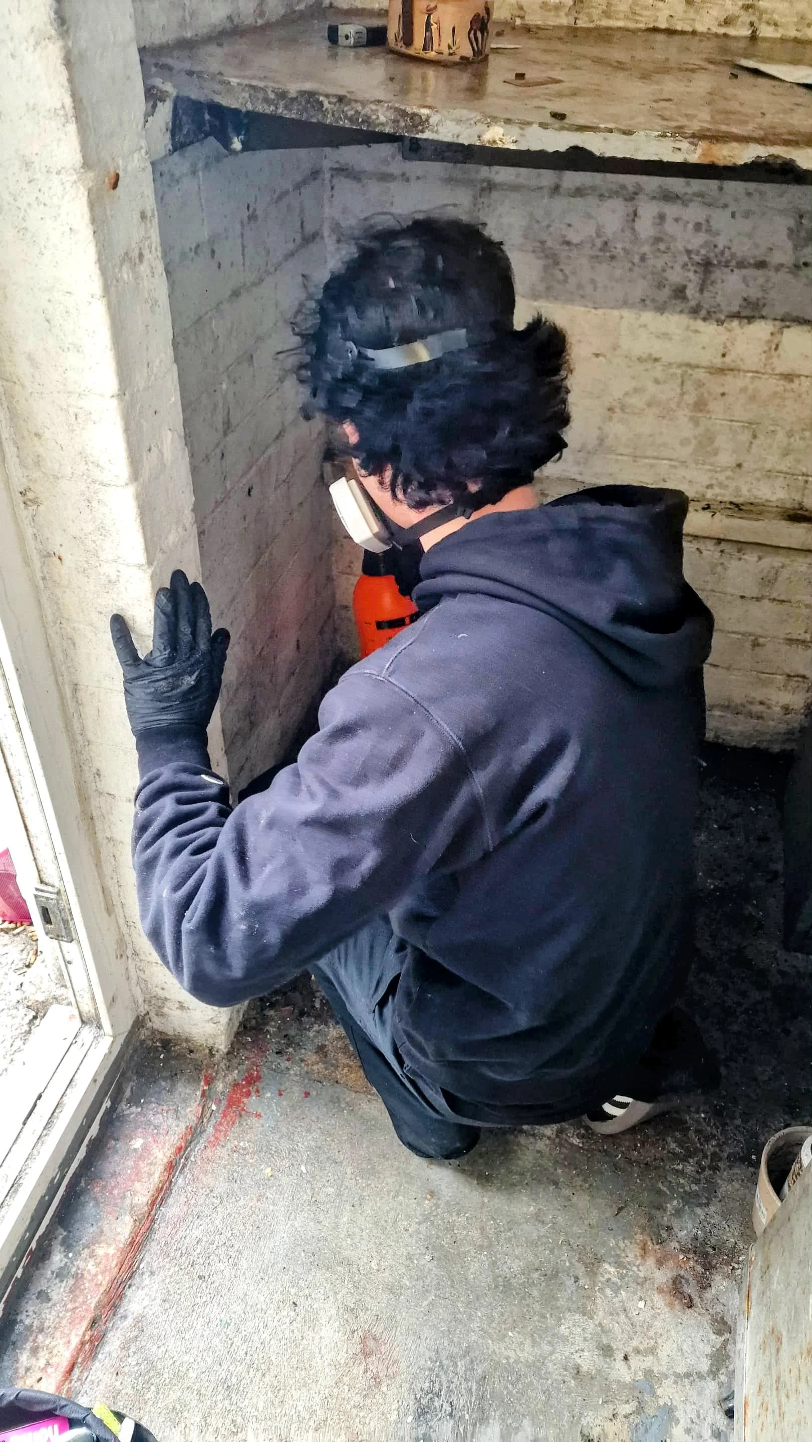 A man kneeling on the ground working on a doorframe or floor area, wearing a dark hoodie, black gloves, a face mask, and glasses. There is construction or renovation material around him.