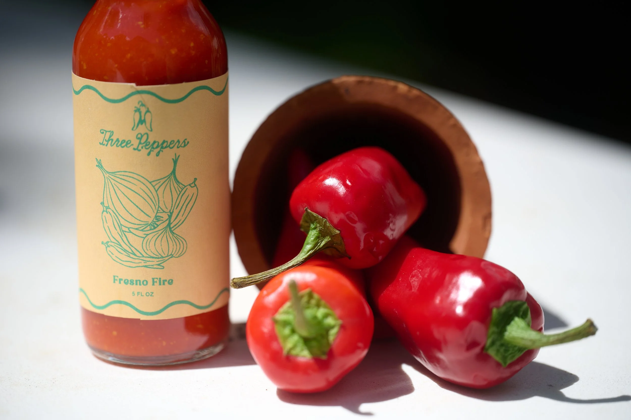 A bottle of Three Peppers Fresno Fire hot sauce surrounded by red peppers on a white surface.