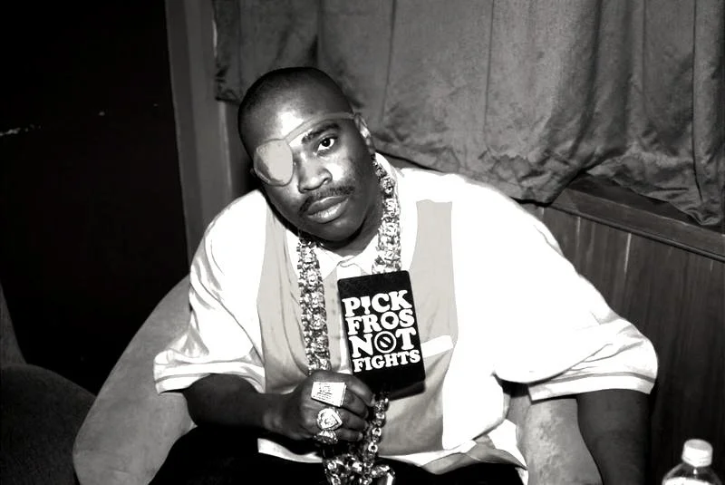 A man wearing an eye patch and holding a microphone with a sign that says 'Pick Fros Not Fights,' sitting on a booth in a dimly lit setting, with curtains behind him.