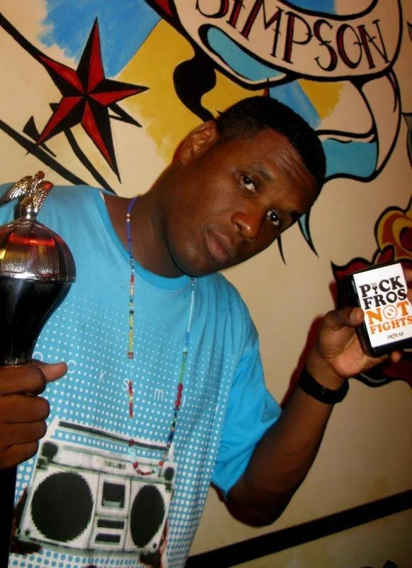 A man with short hair and a blue shirt holding a trophy in one hand and a box with the logo 'Pick Fros Not Fight' in the other, standing in front of a colorful mural with a star and text.