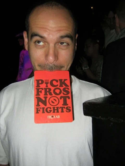 Man holding a red sign that says 'Pick Fros Not Fights' over his mouth at a nightclub or bar.