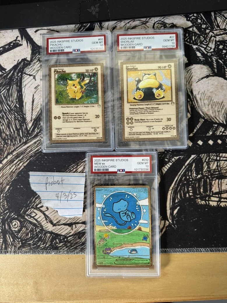 Three vintage Pokémon trading cards in plastic cases, labeled as 2025 INKSPIRATION STUDIOS. The top left card features Pikachu, the top right card features Snorlax, and the bottom card features Mew. The cards are placed on a black and white patterned