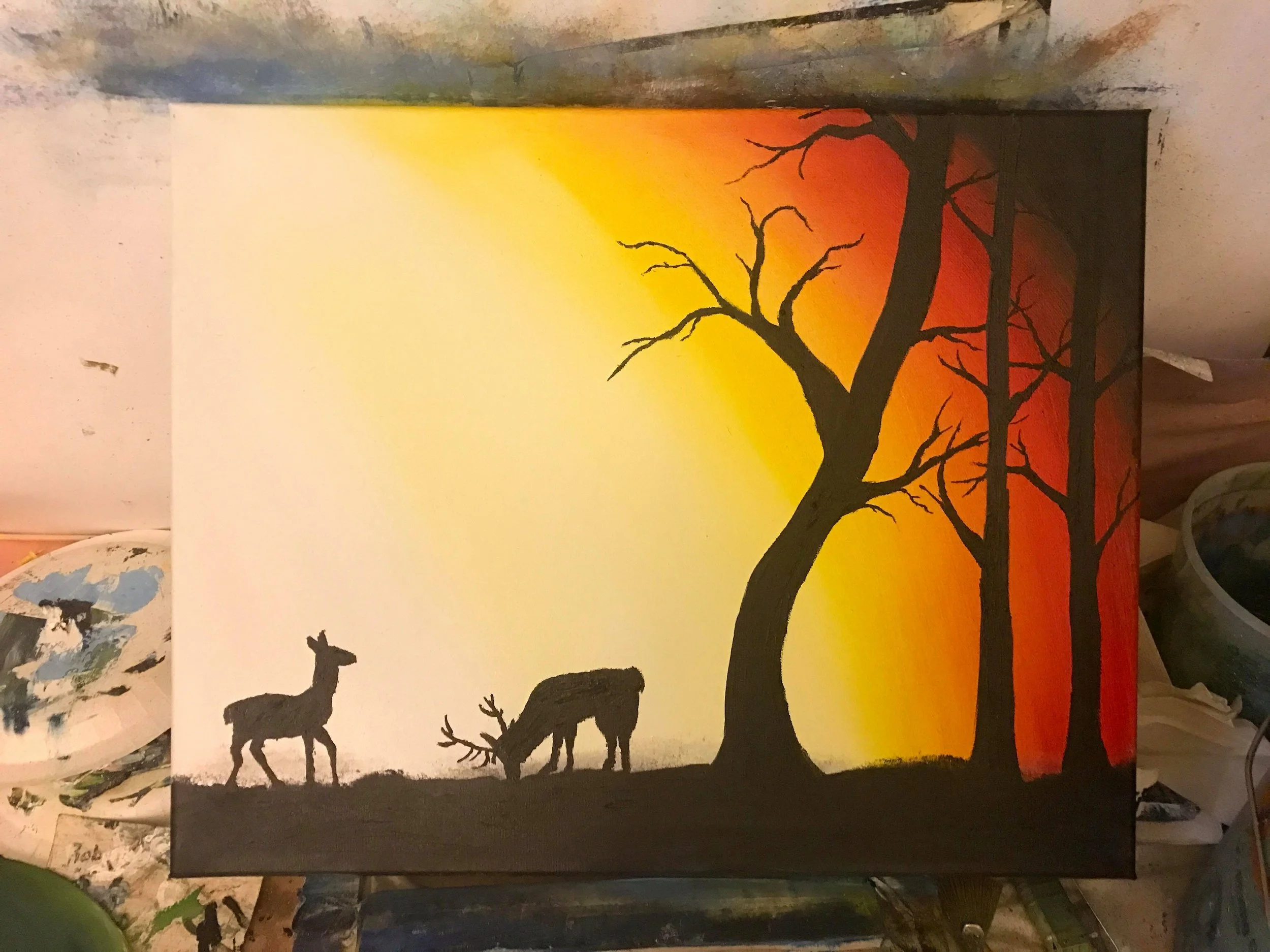 Silhouette of two deer, one standing and one grazing, next to leafless trees against a sunset background with yellow, orange, and red colors.