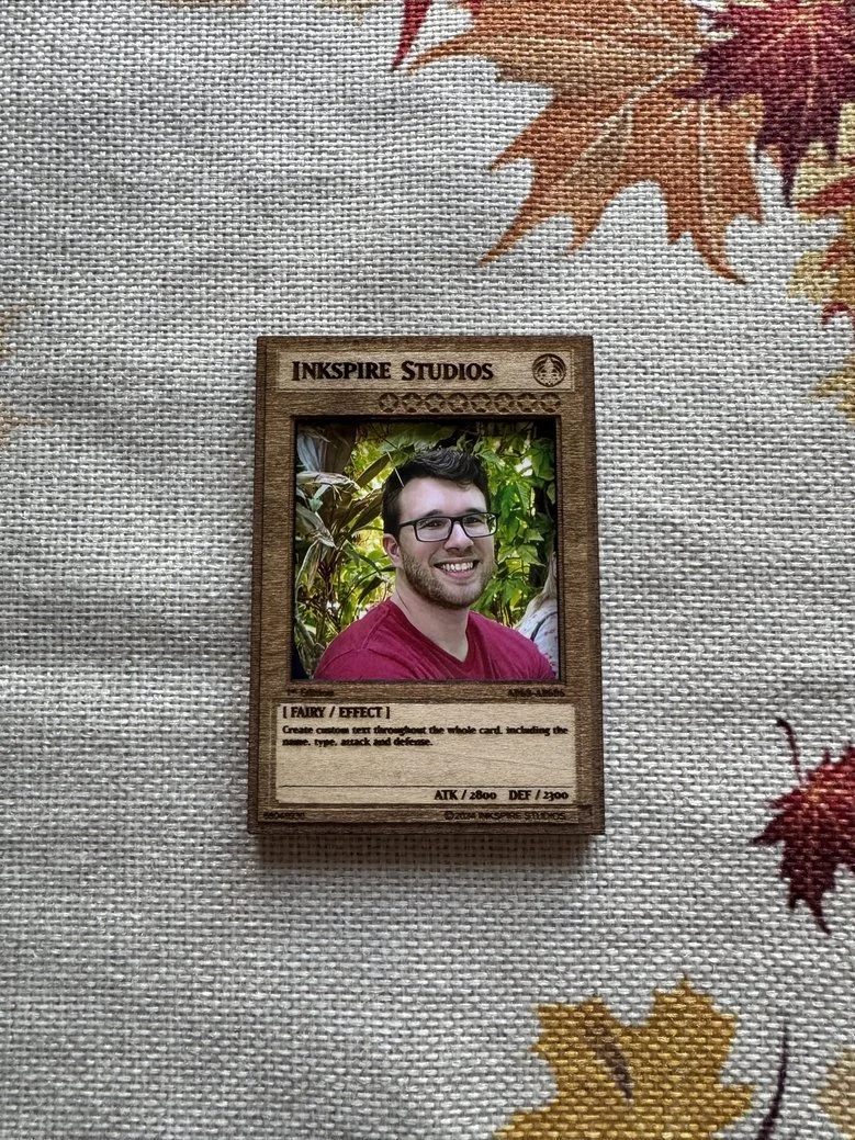 A custom-designed card with a photo of a smiling man wearing glasses and a red shirt. The card is labeled 'Inspire Studios' and has design space for creating text effects. It is placed on a fabric surface with a pattern of fall leaves.