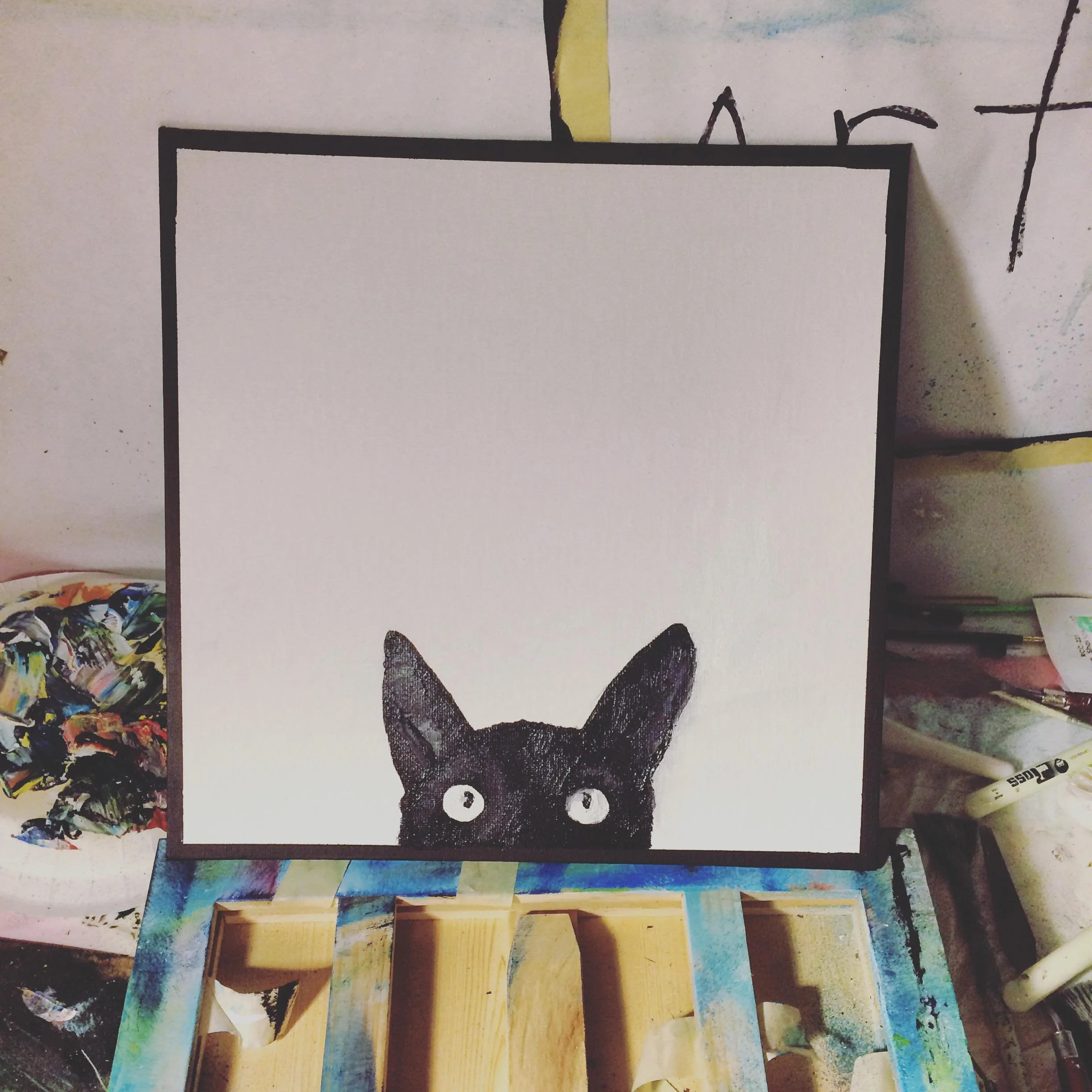 A white canvas with a black cat's head and ears at the bottom, with the cat's eyes made from two buttons, set on a multicolored work table in an art studio.