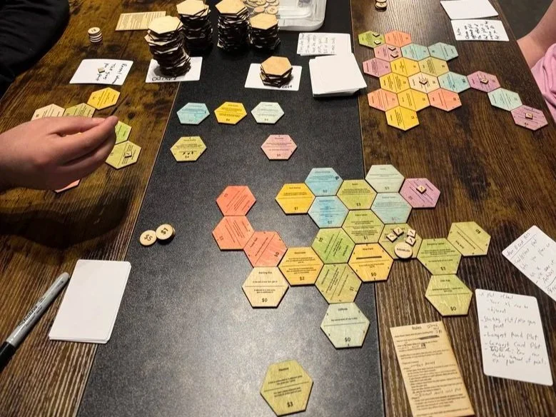 Table with colorful hexagonal tiles used for a board game, along with game pieces, cards, and handwritten notes surrounding it.