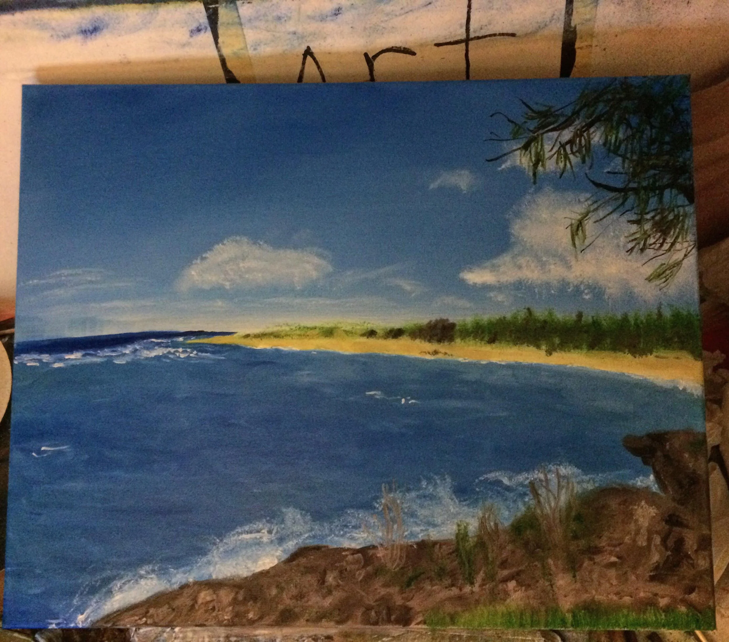 A landscape painting of a coastline with rocky foreground, blue ocean waves, a sandy strip, and a green forest under a partly cloudy sky.