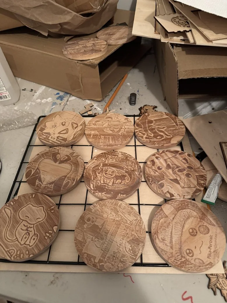 Several wooden coasters with engraved images of Pokémon characters, including Pikachu, Squirtle, Charmander, Bulbasaur, Jigglypuff, and Eevee, arranged on a wire rack in a workshop.