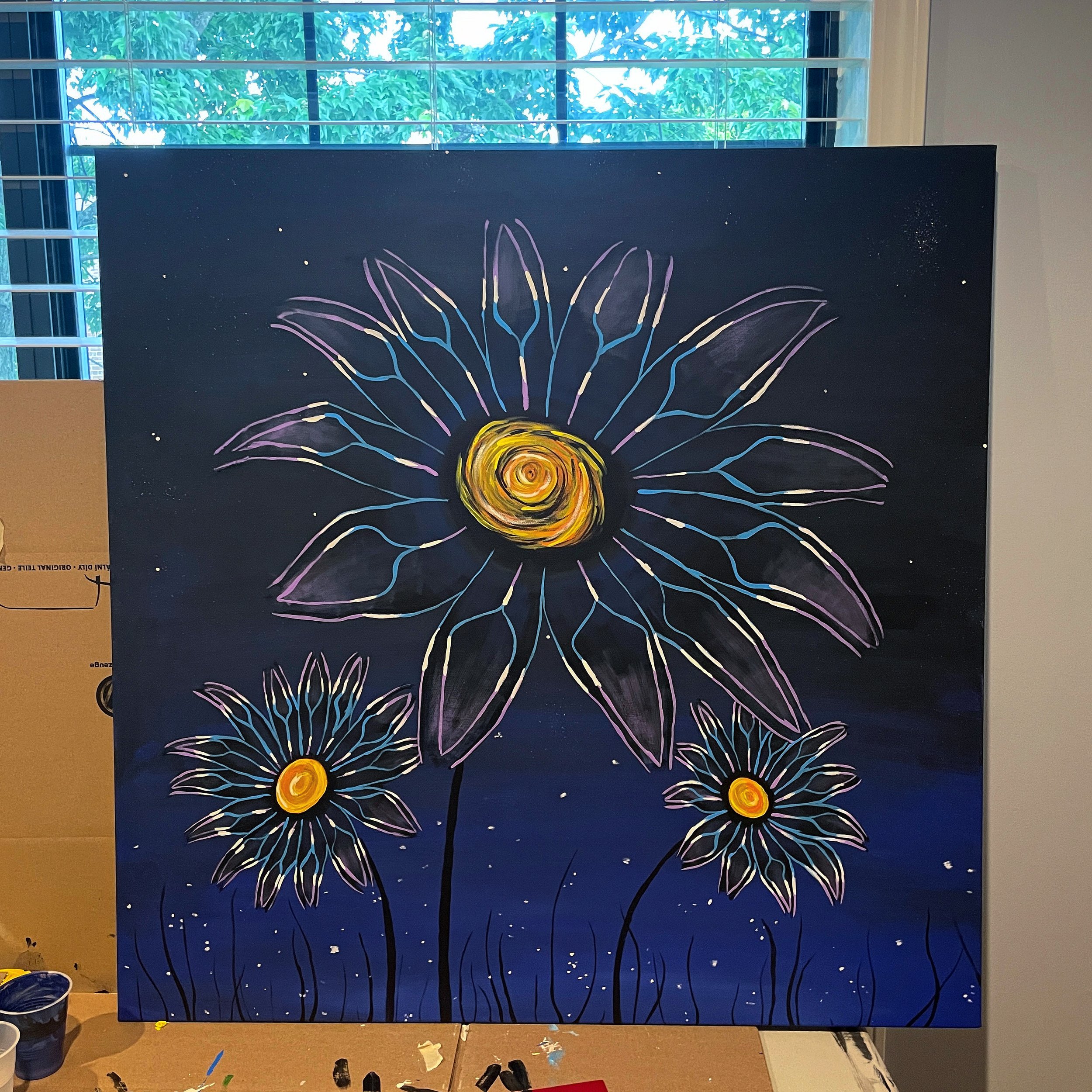 A painting of three large, abstract flowers with yellow centers and multicolored petals on a dark blue background, with a starry night sky and green trees visible through a window behind the painting.