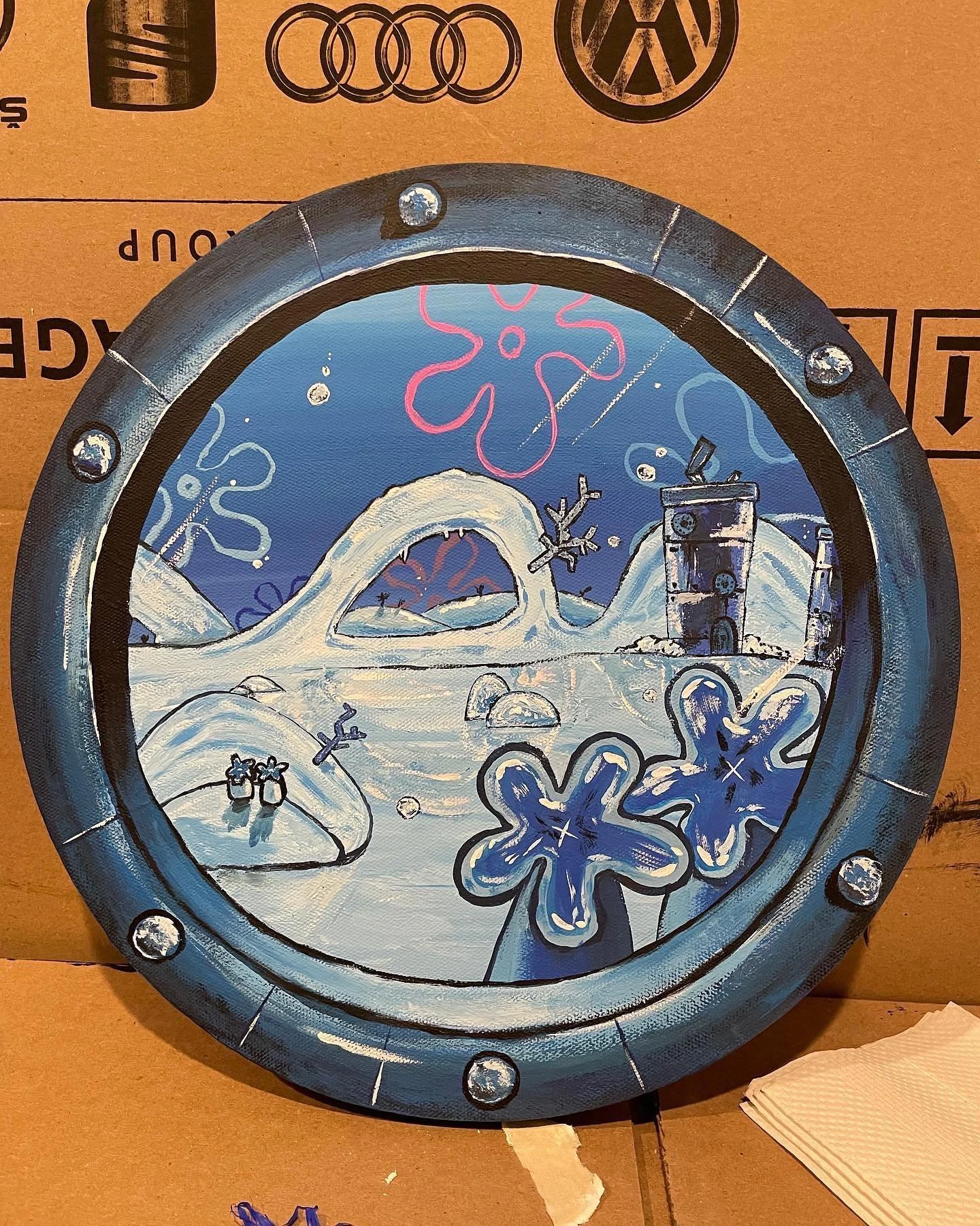 A decorative round sign featuring a winter scene with snow-covered hills, an icy arch, snowflakes, and a small building, painted in shades of blue, white, pink, and black.