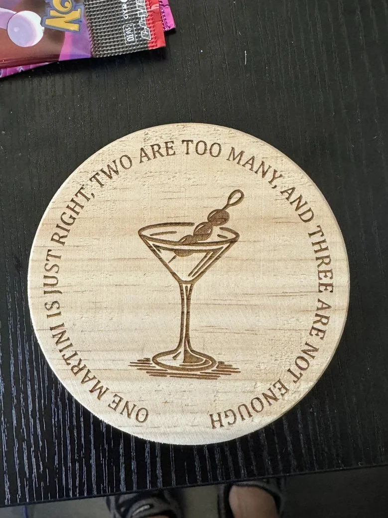 Wooden coaster with engraved quote and drawing of a martini glass with an olive on a toothpick. The quote reads, 'ONE MARTINI IS JUST RIGHT, TWO ARE TOO MANY, AND THREE ARE NOT ENOUGH.'