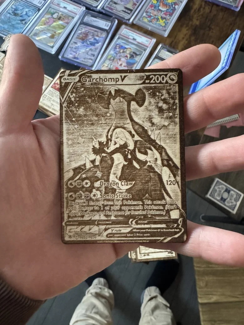 A person holding a holographic Pokémon card featuring Garchomp V, with a background of various Pokémon cards in protective sleeves on a table.