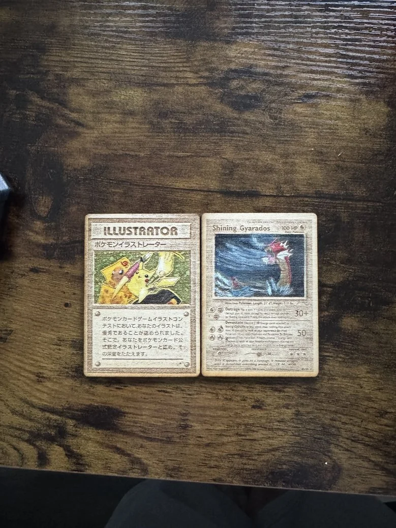 Two vintage Pokémon cards on a wooden surface. The card on the left features Pikachu with the title 'Illustrator' and Japanese text, while the card on the right features Gyarados titled 'Shining Gyarados' with Japanese text and artwork of the Pokémon