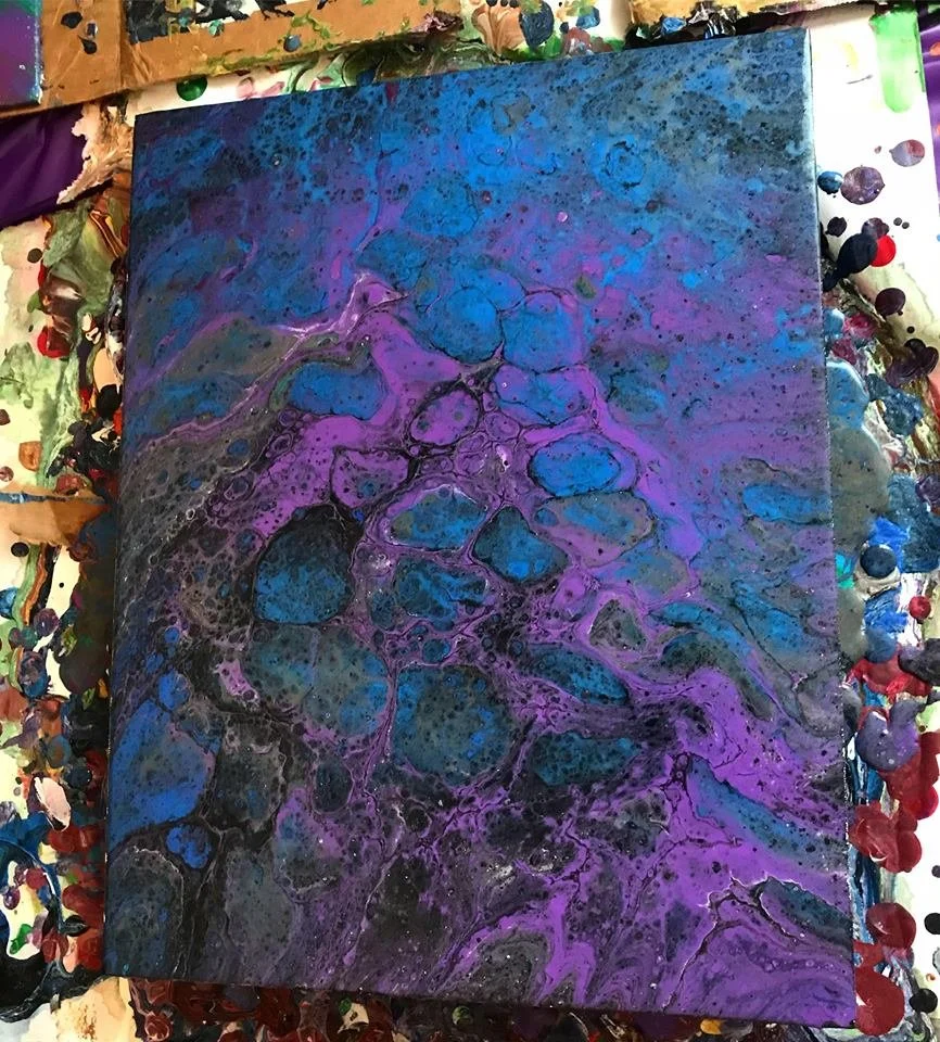 Abstract painting with blue, purple, and black marbled swirls on a canvas, surrounded by paint splatters and palette remnants.
