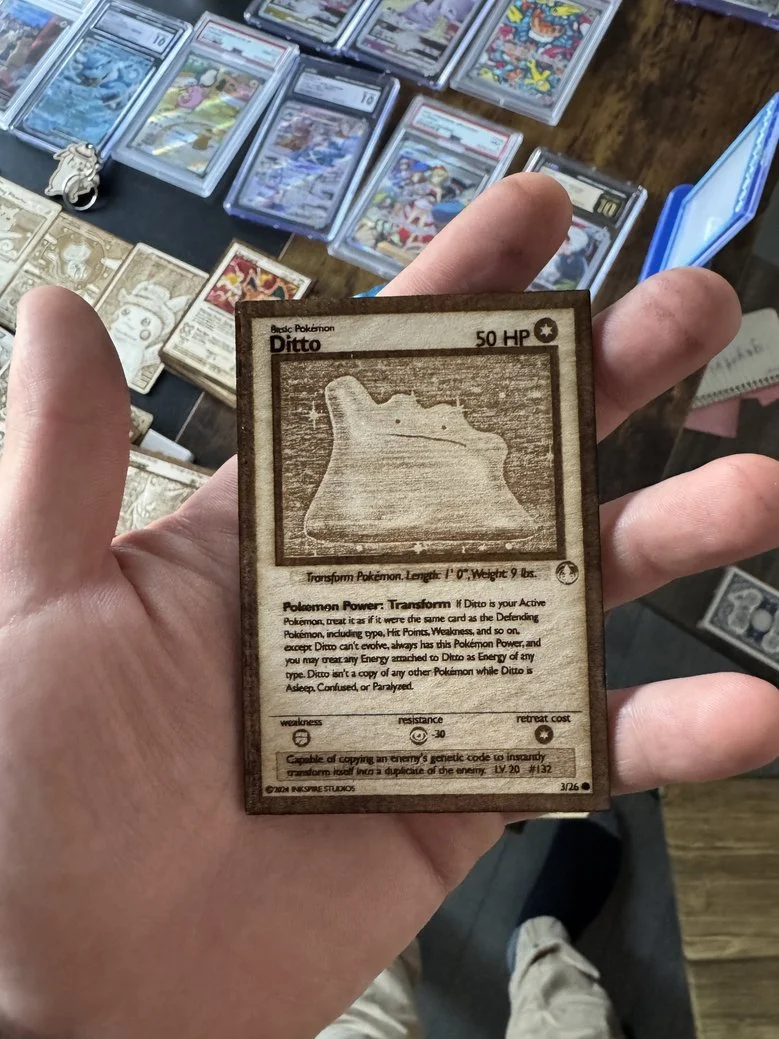 Person holding a Pokémon card for Ditto with 50 HP, showing its map silhouette, against a background of various trading cards on a table.