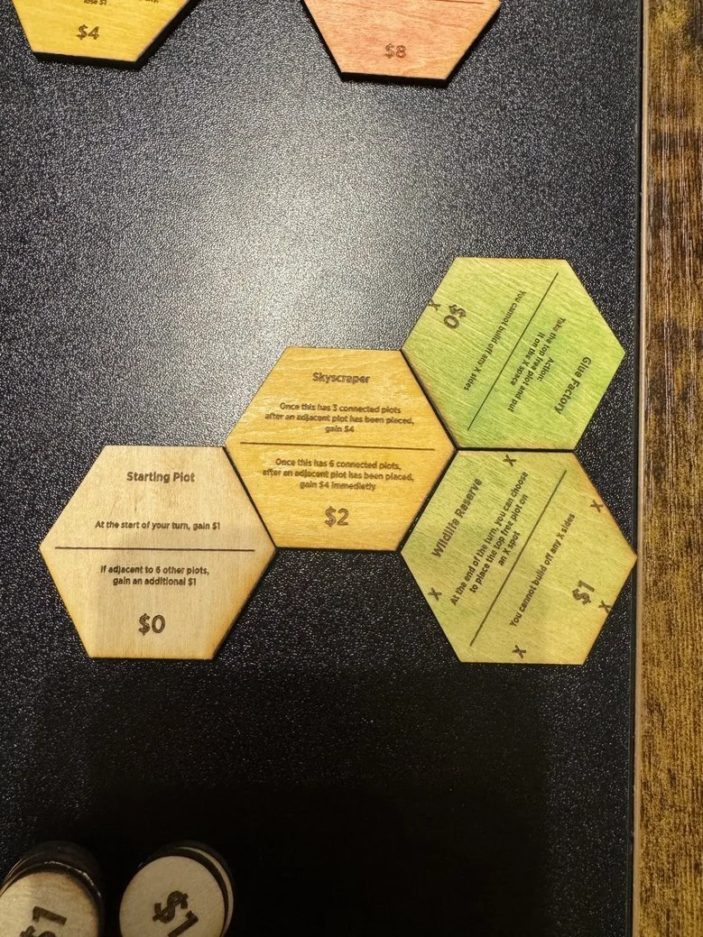 Game pieces on a dark tabletop for a board game involving hexagonal tiles with instructions and point values, including a starting plot and connected plots, in a casual setting.