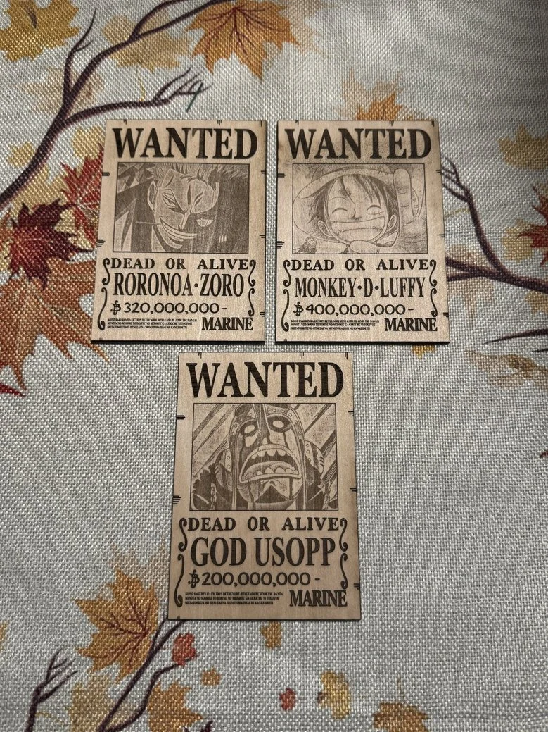 Three illustrated 'wanted' posters on a fabric surface with autumn leaves pattern. The posters feature characters from the anime One Piece, including Roronoa Zoro, Monkey D. Luffy, and God Usopp, with their bounty amounts.