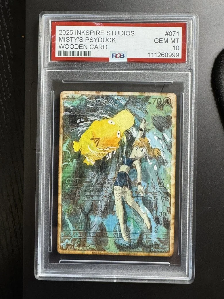 A trading card inside a clear plastic case with a red and white label. The card features a cartoon girl with short hair, wearing a dark blue outfit, and a large yellow fish with big eyes in an aquatic scene.