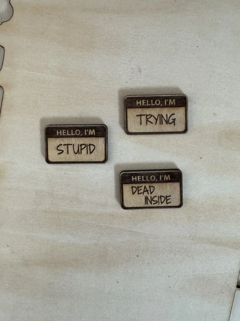 Three black and white name tags with handwritten text that read 'Hello, I'm Stupid', 'Hello, I'm Trying', and 'Hello, I'm Dead Inside' on a light-colored surface.