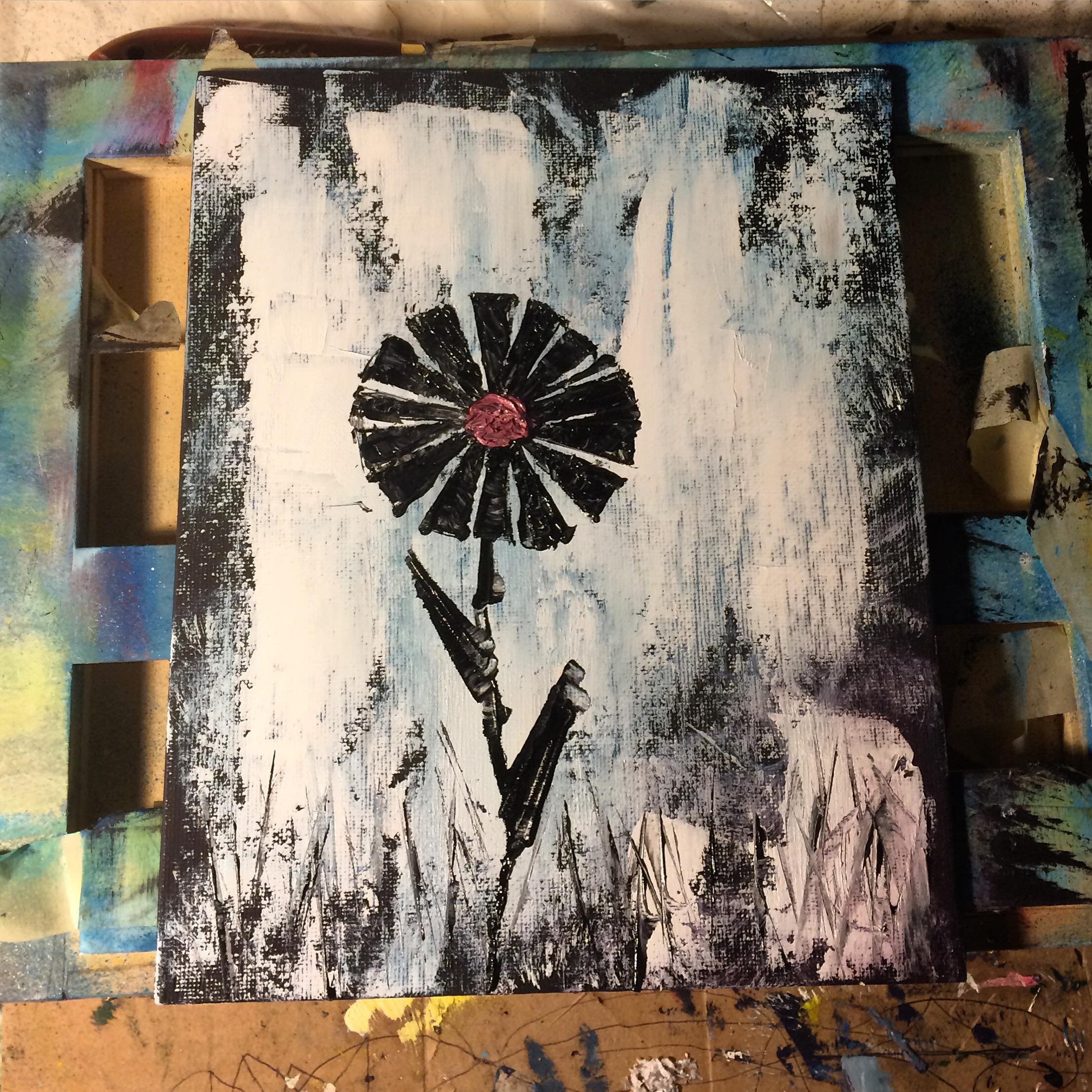 Abstract painting of a black flower with a pink center on a textured background of black and white. The painting appears to be on a canvas resting on a colorful, splattered art table.