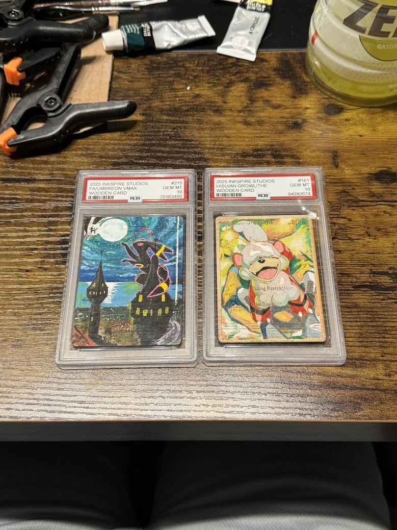 Two graded Pokémon trading cards in clear cases on a wooden table. The left card features a dark silhouette of a Pokémon with a moon and a spooky castle in the background, labeled as 2025 INKSPRE STUDIOS FAUXMBREM VMAX WOODEN CARD. The right card dep