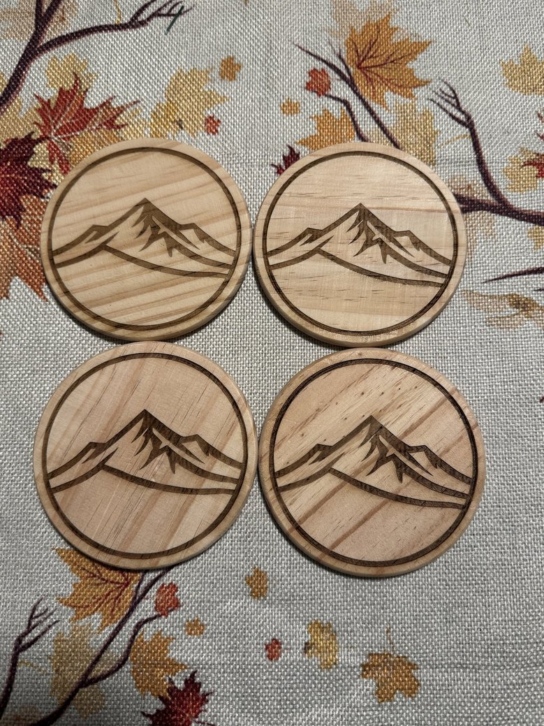Four round wooden coasters with engraved mountain designs, placed on a tablecloth with autumn leaves pattern.