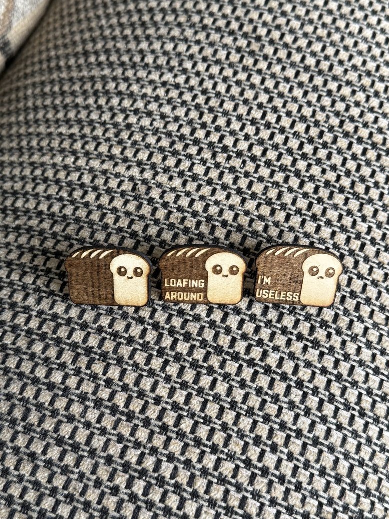 Three wooden pins with cute ghost face designs and humorous text, placed on a textured fabric surface. The pins say, from left to right, 'LOAFING AROUND,' 'LOAFING AROUND,' and 'I'M USELESS.'