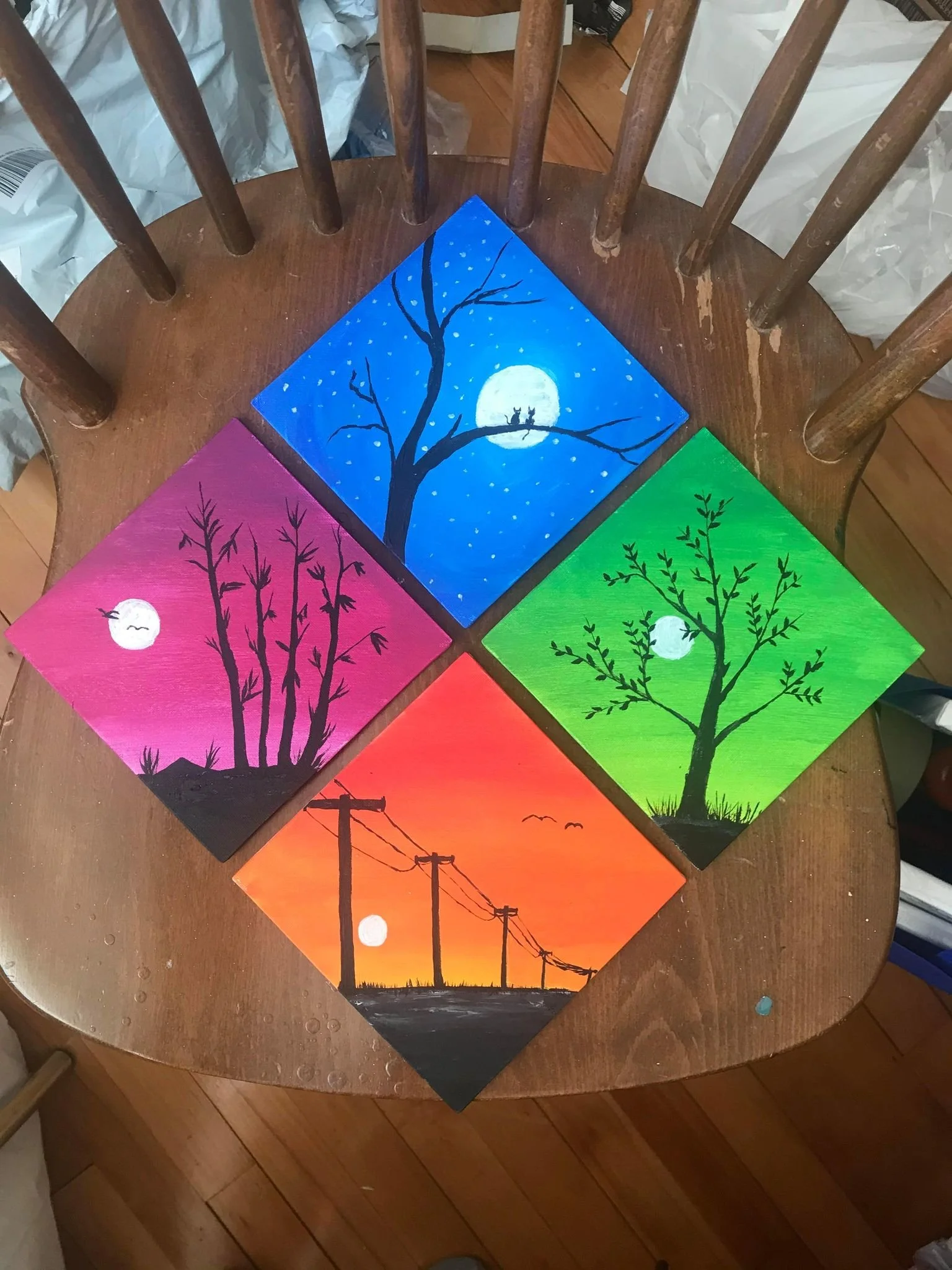 Four colorful paintings of different trees and outdoor scenes arranged in a square on a wooden chair.