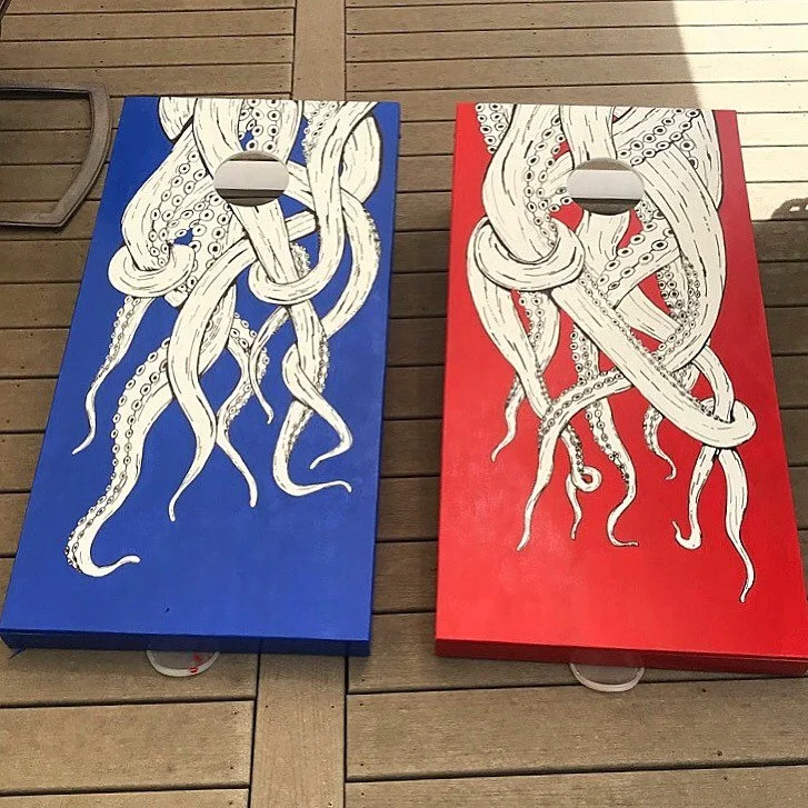 Two cornhole boards with octopus artwork, one blue and one red, placed on a wooden deck.