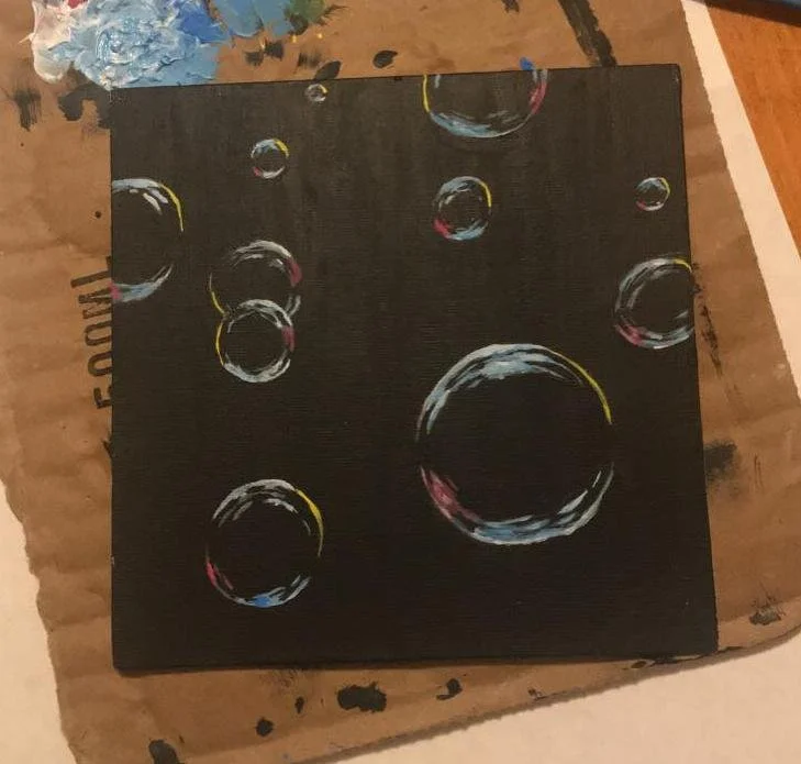 A black canvas with painted bubbles of various sizes, accented with hints of yellow, pink, and blue, resting on a wooden surface.