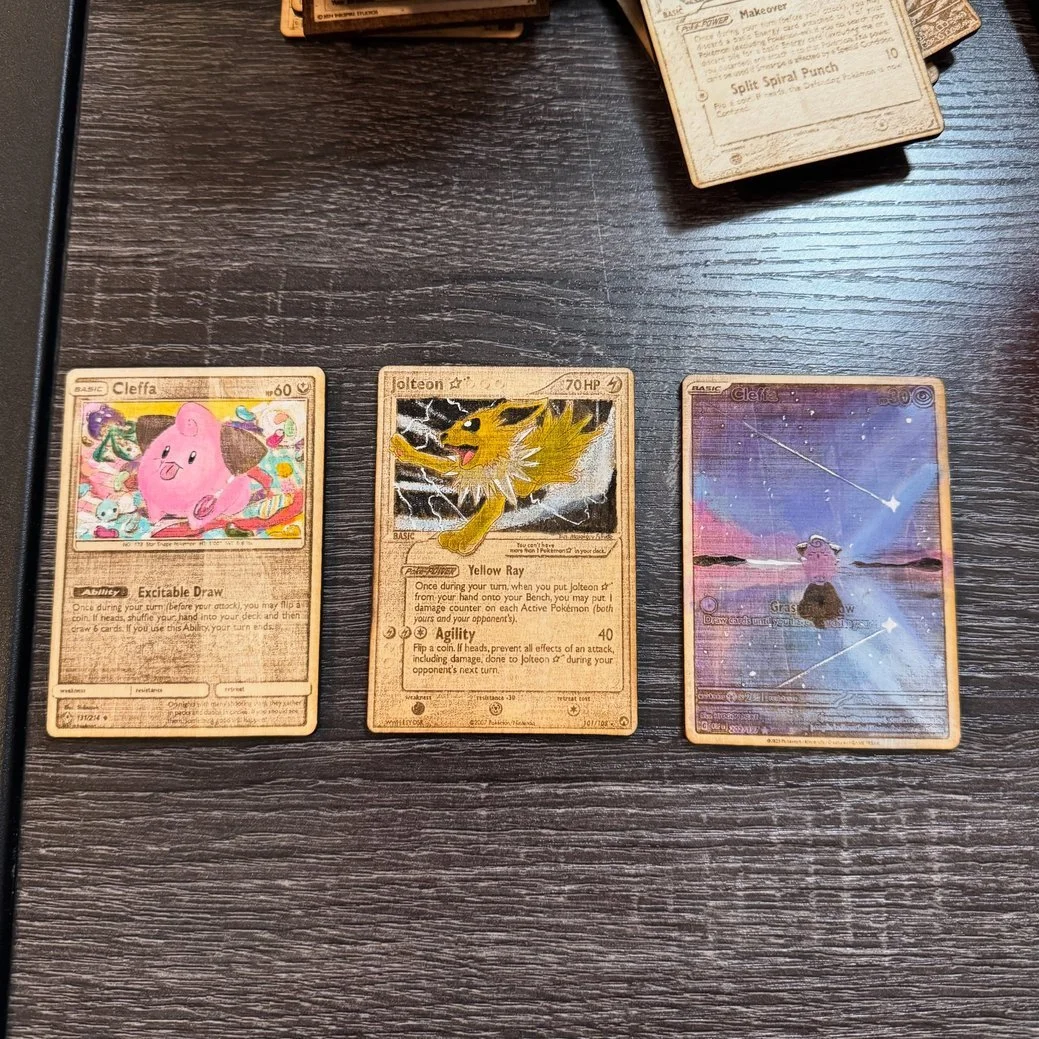 Three Pokémon trading cards laid out flat on a dark wooden surface, with additional cards partially visible at the top right corner. The cards feature Cleffa, Jolteon, and a Cleffa with a starry background.
