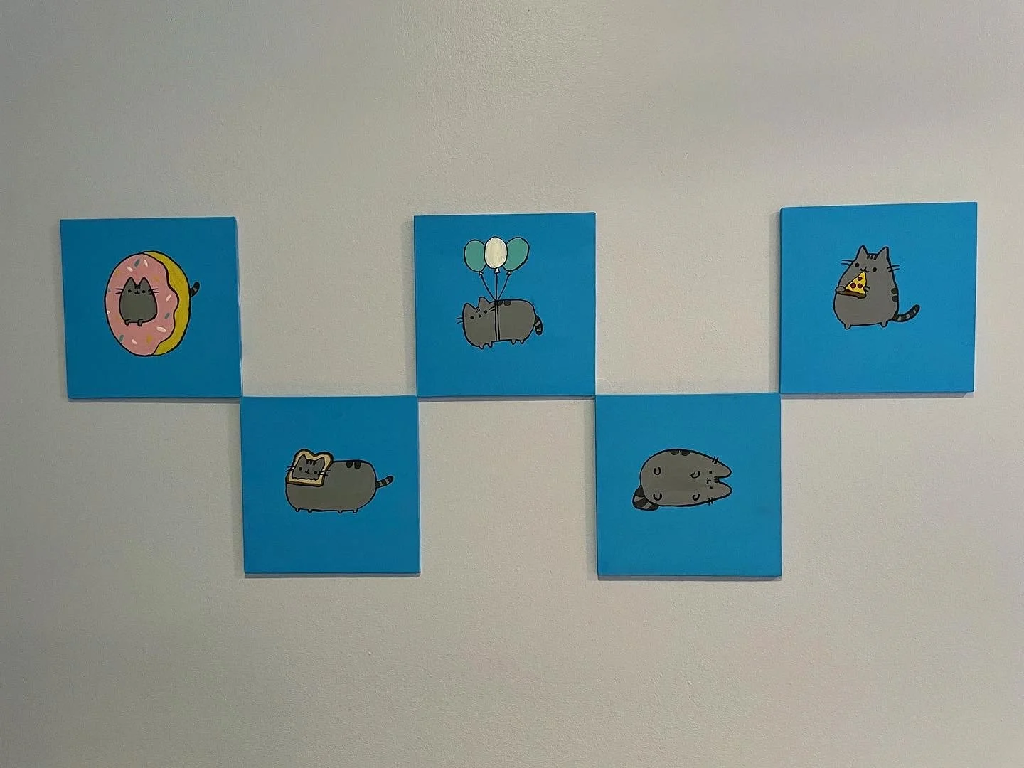 Six small blue canvases with cartoon cat illustrations arranged on a white wall, forming a horizontal cross pattern. The illustrations depict cats with various accessories and in different poses.
