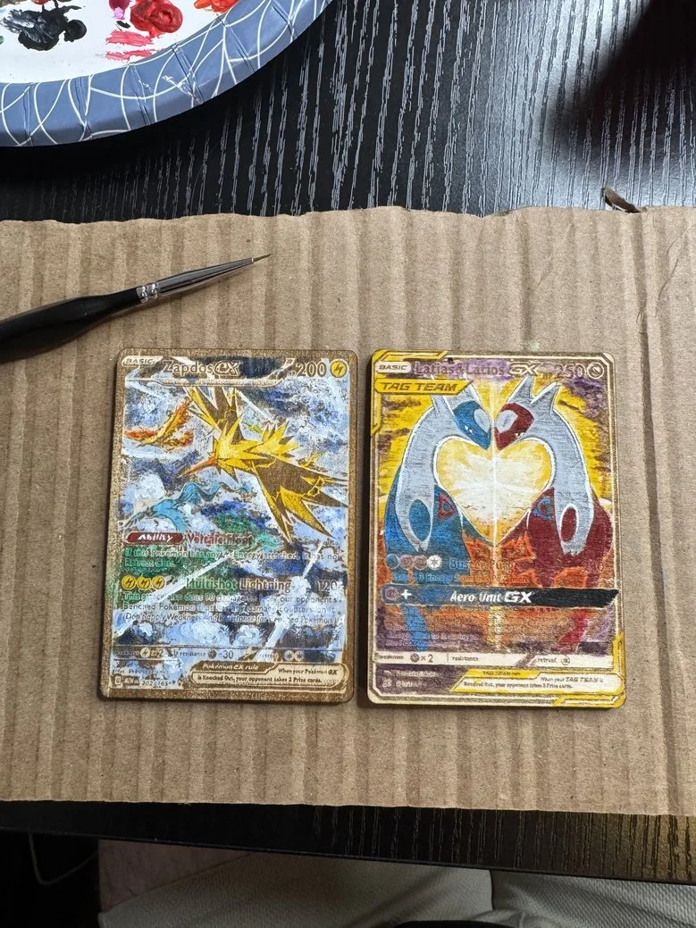 Two Pokémon trading cards on a piece of cardboard, one featuring Zapdos GX and the other featuring a Tag Team Gardevoir and Gardevoir GX, with a pen and a decorative plate partially visible in the background.