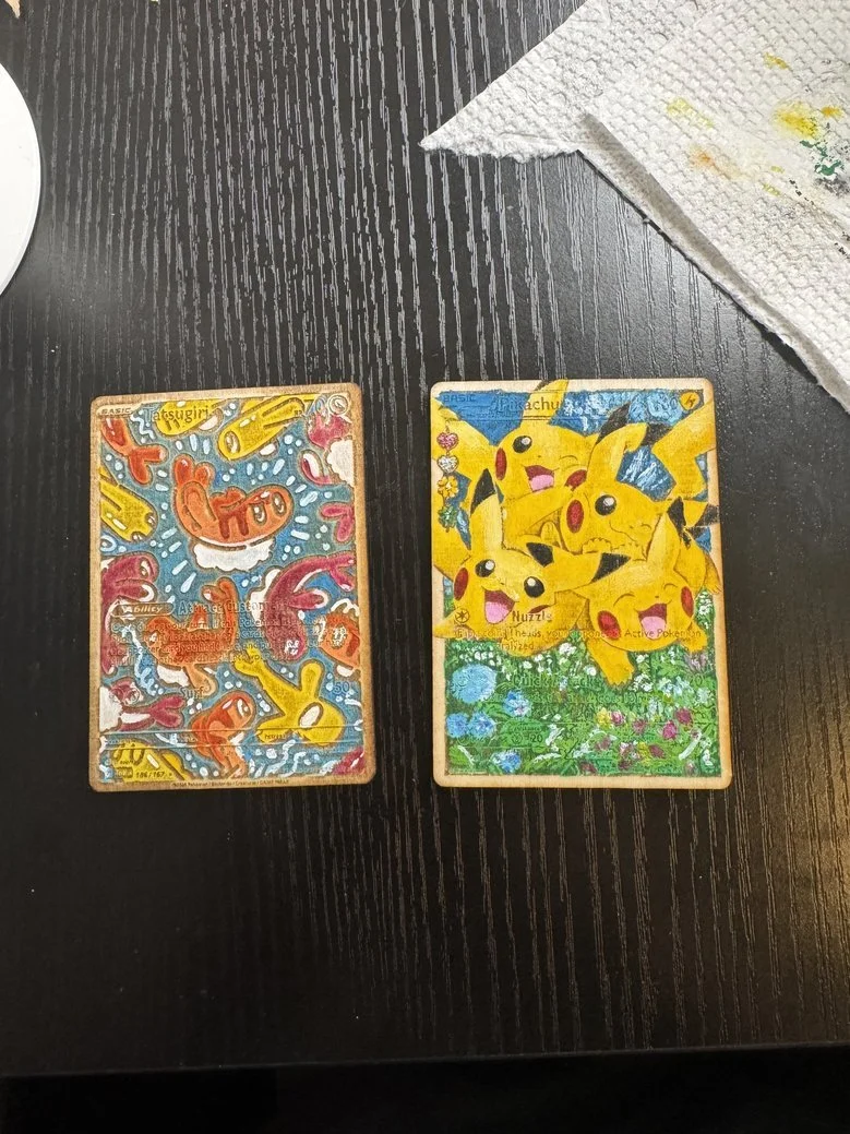 Two Pokémon cards on a dark wooden surface, one featuring Ditto and the other featuring Pikachu with multiple Pikachu illustrations, alongside a white paper towel with colored stains.